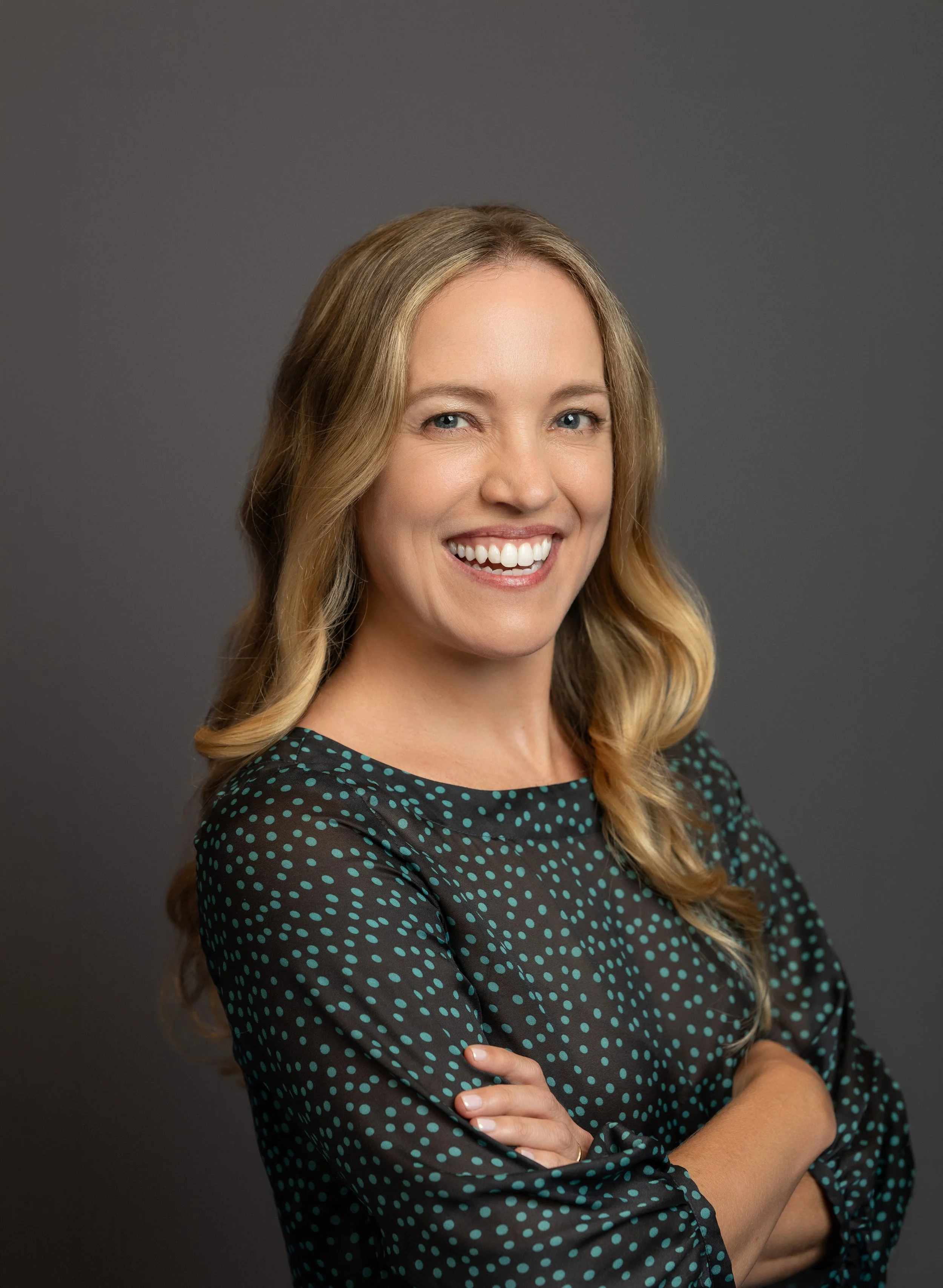 Natalie Rella, the Communications Specialist at the UF Center for Arts in Medicine, standing with crossed arms, wearing a black dress with teal polka dots, standing against a dark-gray background.
