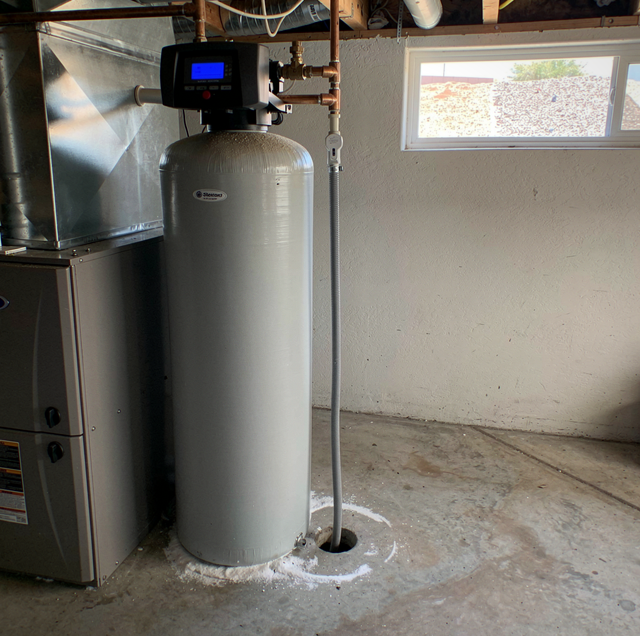 A tall cylindrical metal water tank with digital control panel in a basement next to furnace equipment and piping.