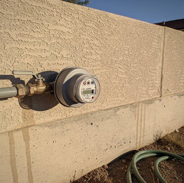 Water meter mounted on a textured stucco wall with pipes and valve attached.