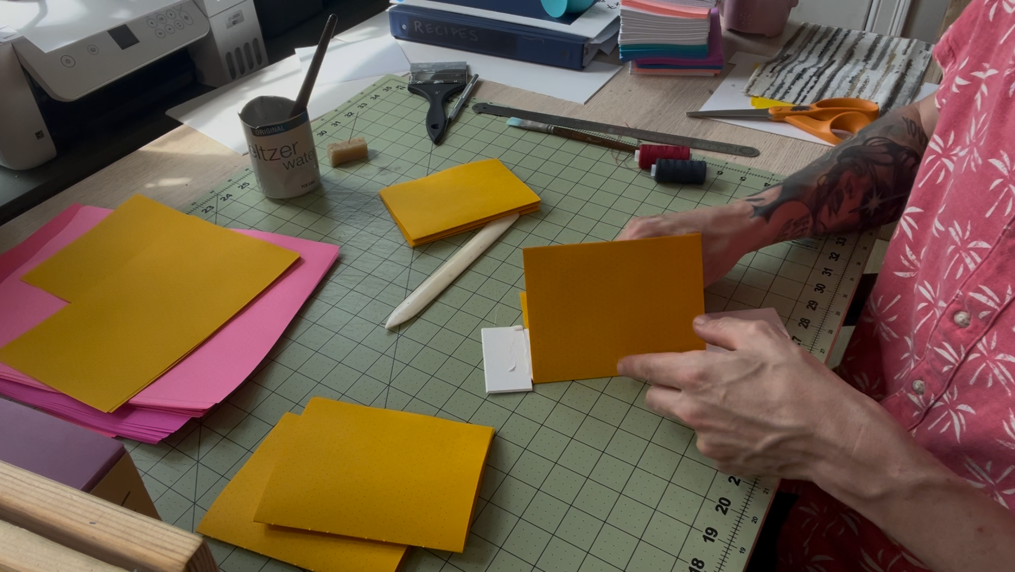 Person with tattoos folding yellow paper on a craft cutting mat surrounded by stacks of pink and yellow paper, scissors, scissors, and workspace supplies.