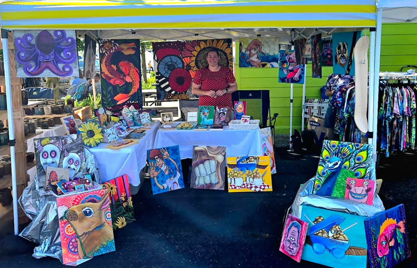 Art booth at outdoor market featuring colorful paintings of animals, fantasy characters, and abstract designs, with a woman in a red polka dot dress standing behind the display.