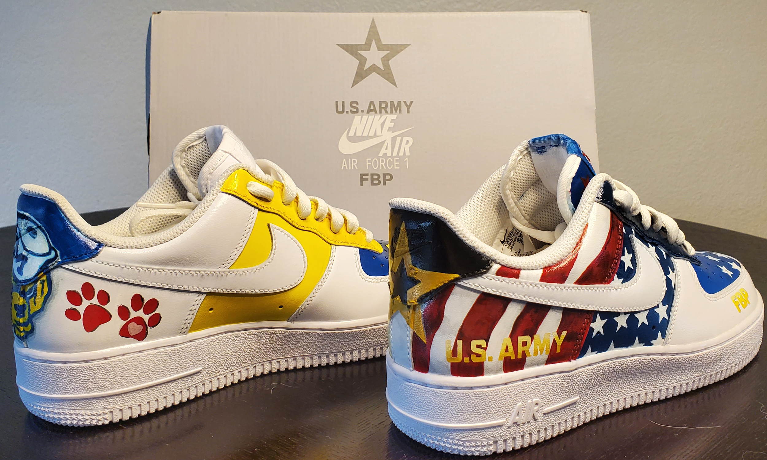 Pair of customized Nike Air Force 1 sneakers with U.S. Army themes, featuring stars and stripes and military-inspired graphics, placed in front of a white U.S. Army Nike Air Force 1 box.