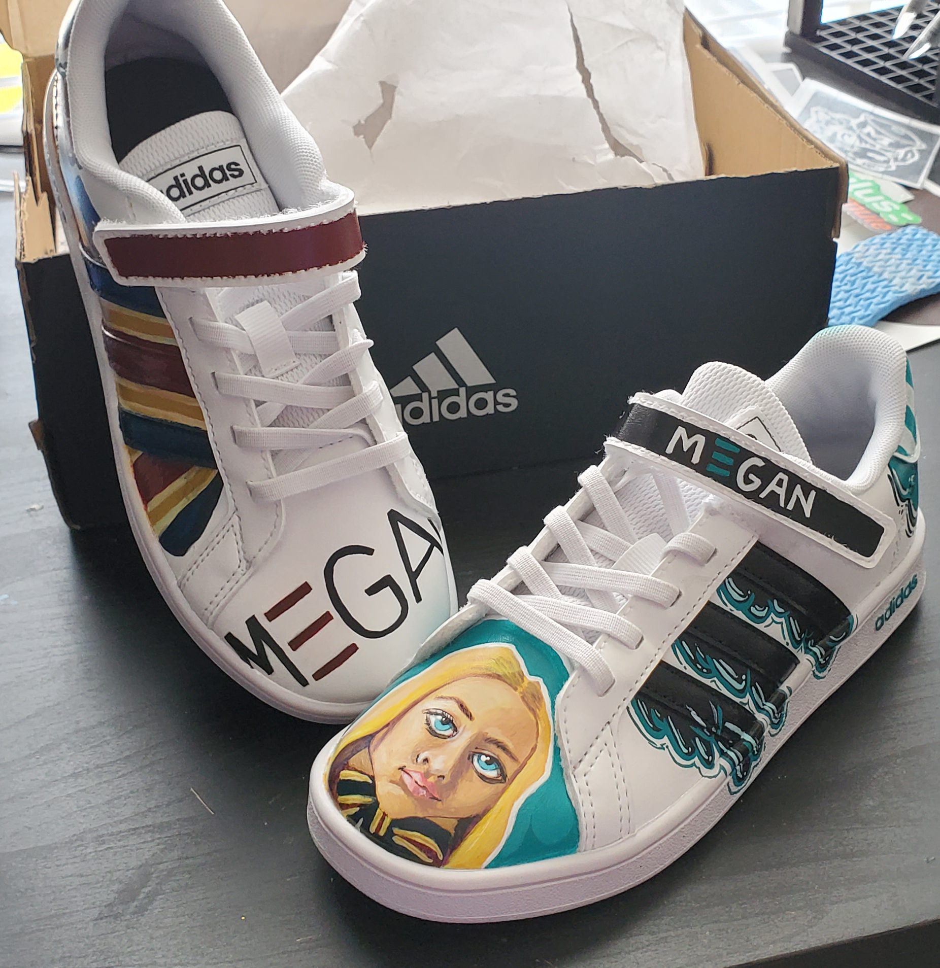 White sneakers with rainbow stripes and a portrait of a woman painted on the side, placed on a black surface with a black Adidas shoe box in the background.