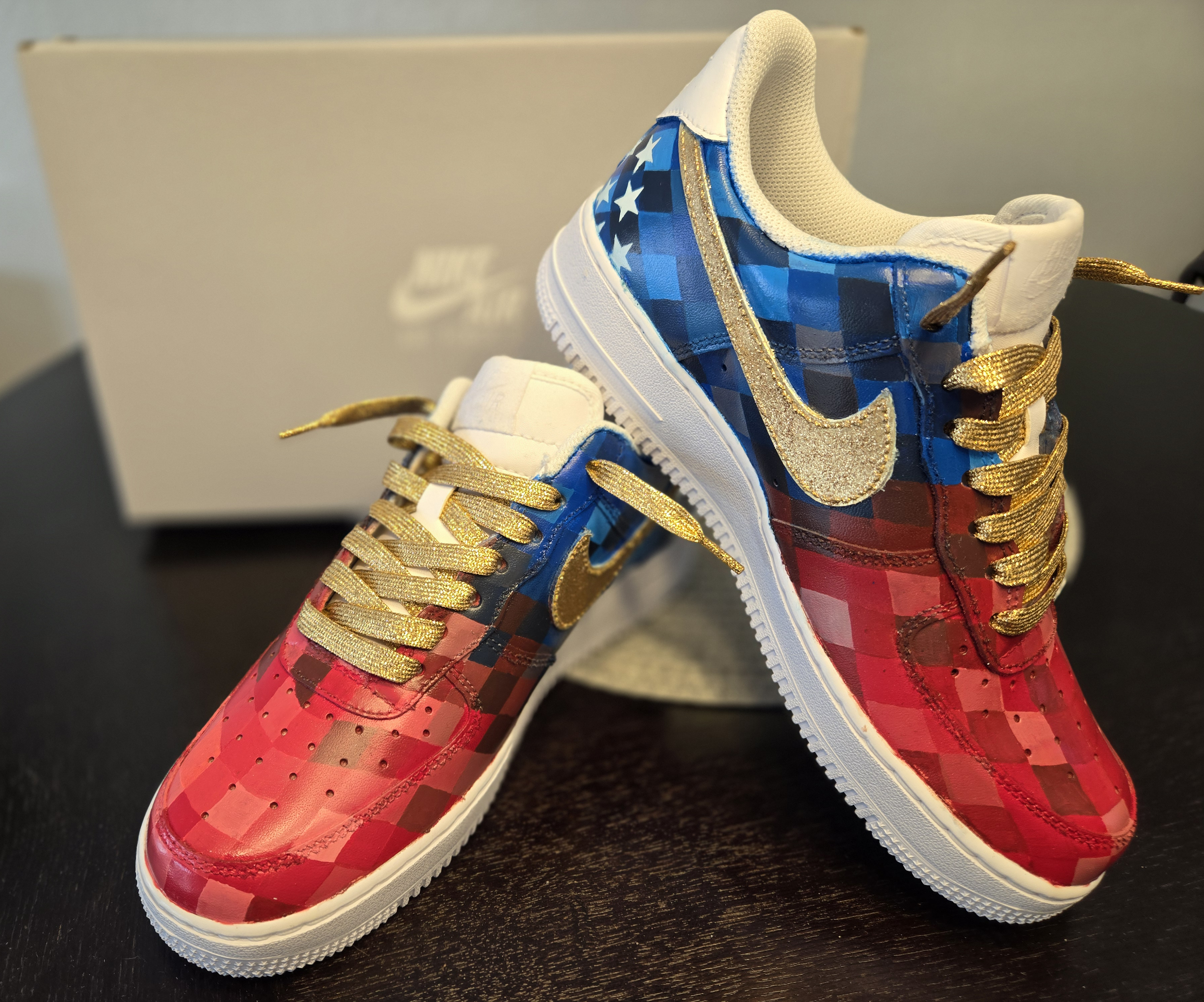 Close-up of a pair of colorful sneakers, one red and one blue, with gold laces and gold accents, on a black surface with a beige box in the background.