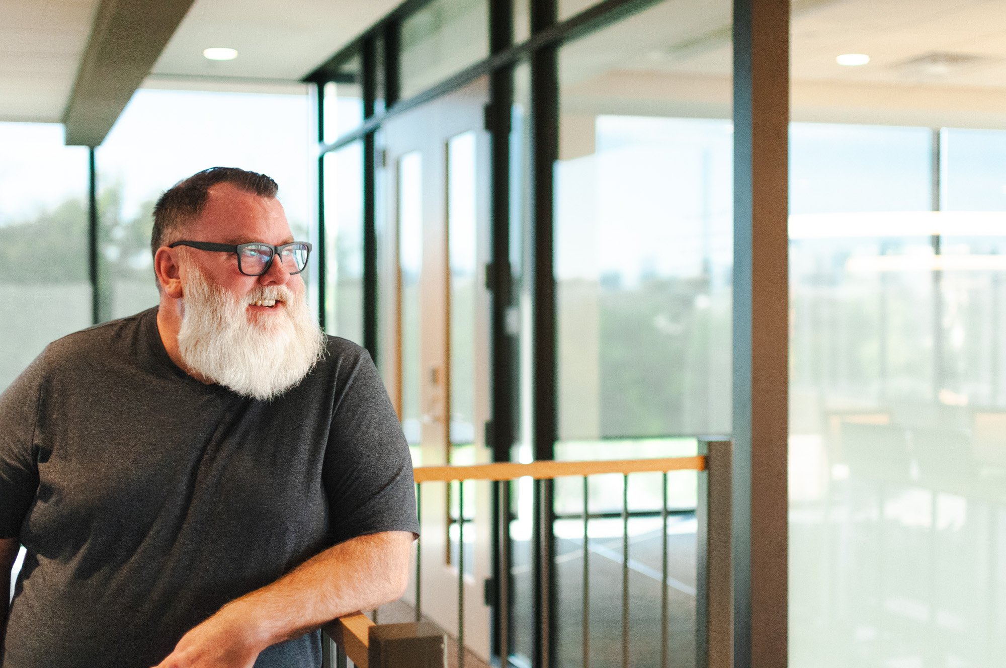 A smiling man with glasses, a beard, and a mustache, wearing a dark t-shirt, sitting indoors near large windows with natural light.