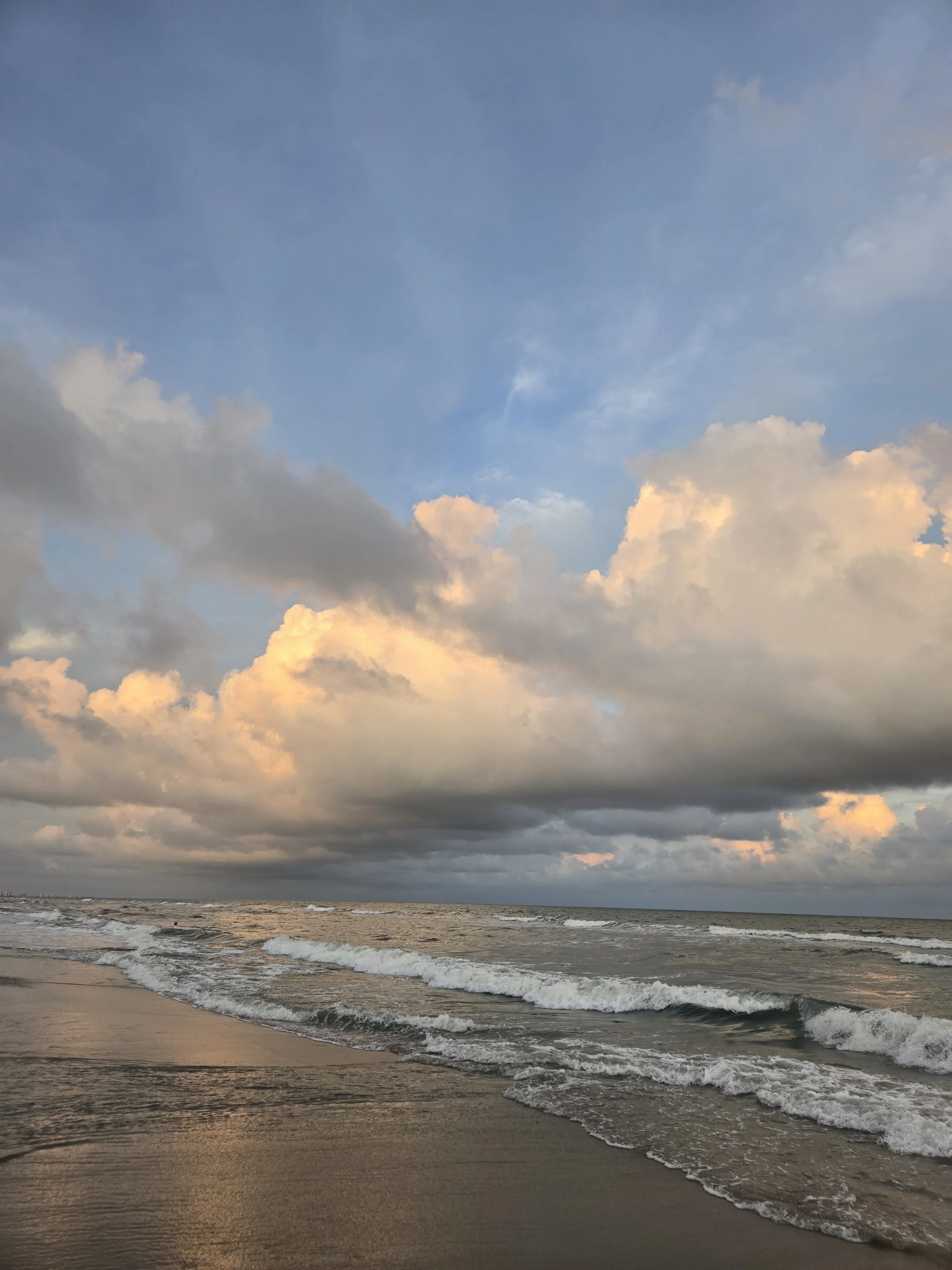 A beach scene during sunset with clouds in a mostly cloudy sky and gentle waves crashing onto the shoreline.