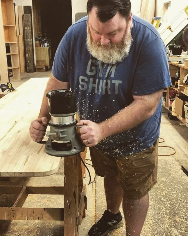 Man with a beard using a router on a wooden workbench in a woodworking shop.