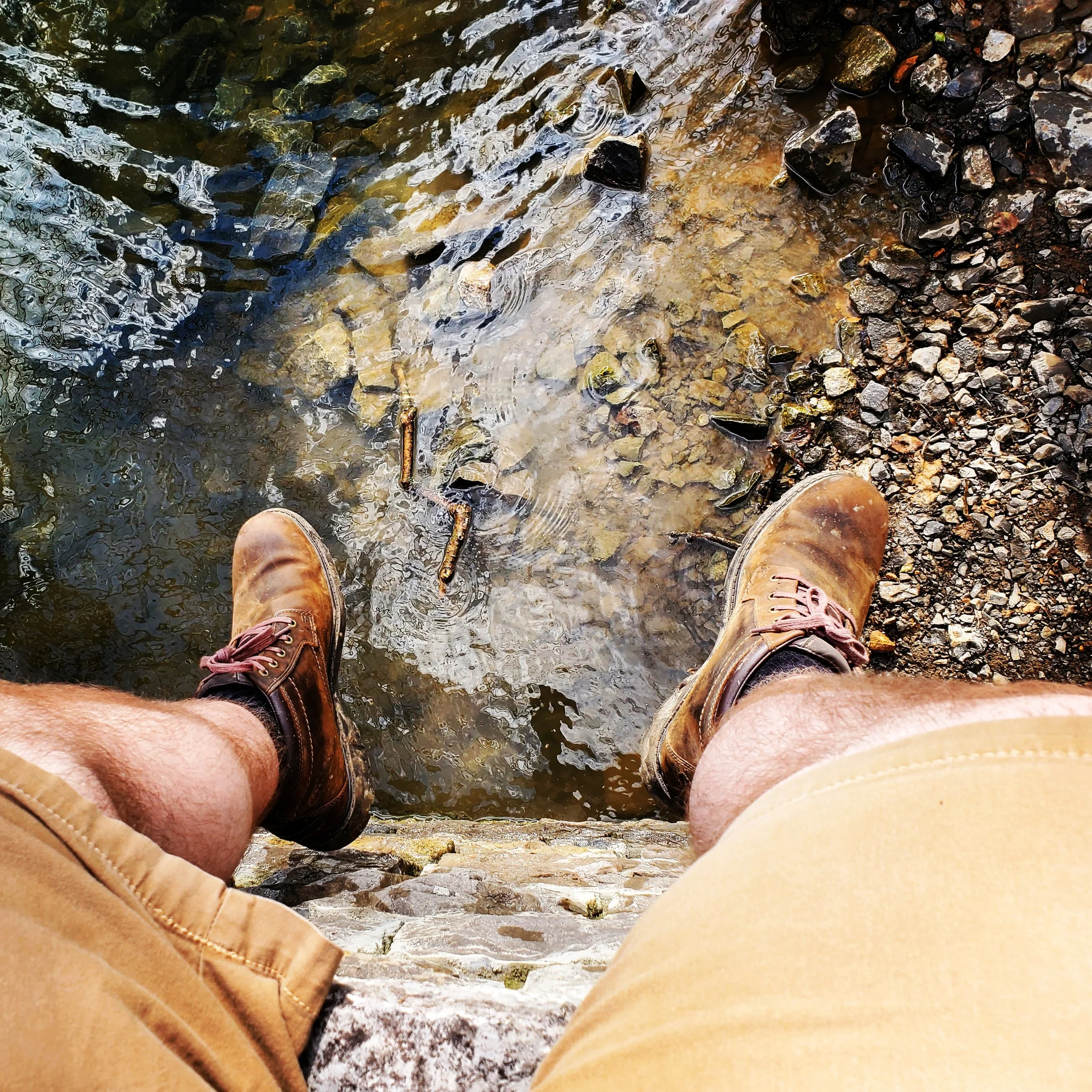 Person standing on rocky ground near a river or creek, with water flowing over rocks and pebbles, wearing brown boots and tan shorts.