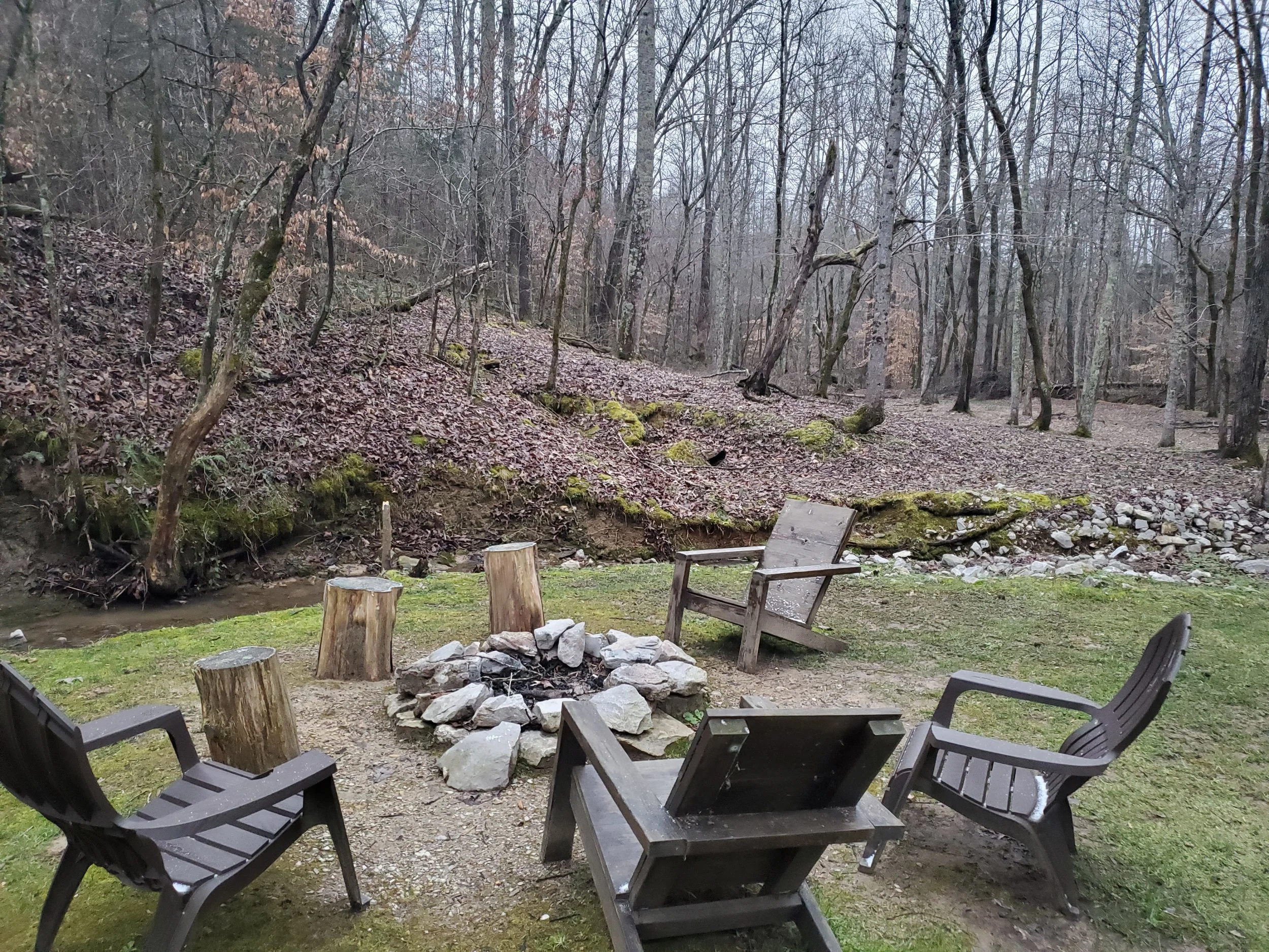 A backyard scene with a fire pit surrounded by five black plastic chairs, on a grassy area near a creek, with leafless trees on a cloudy day.