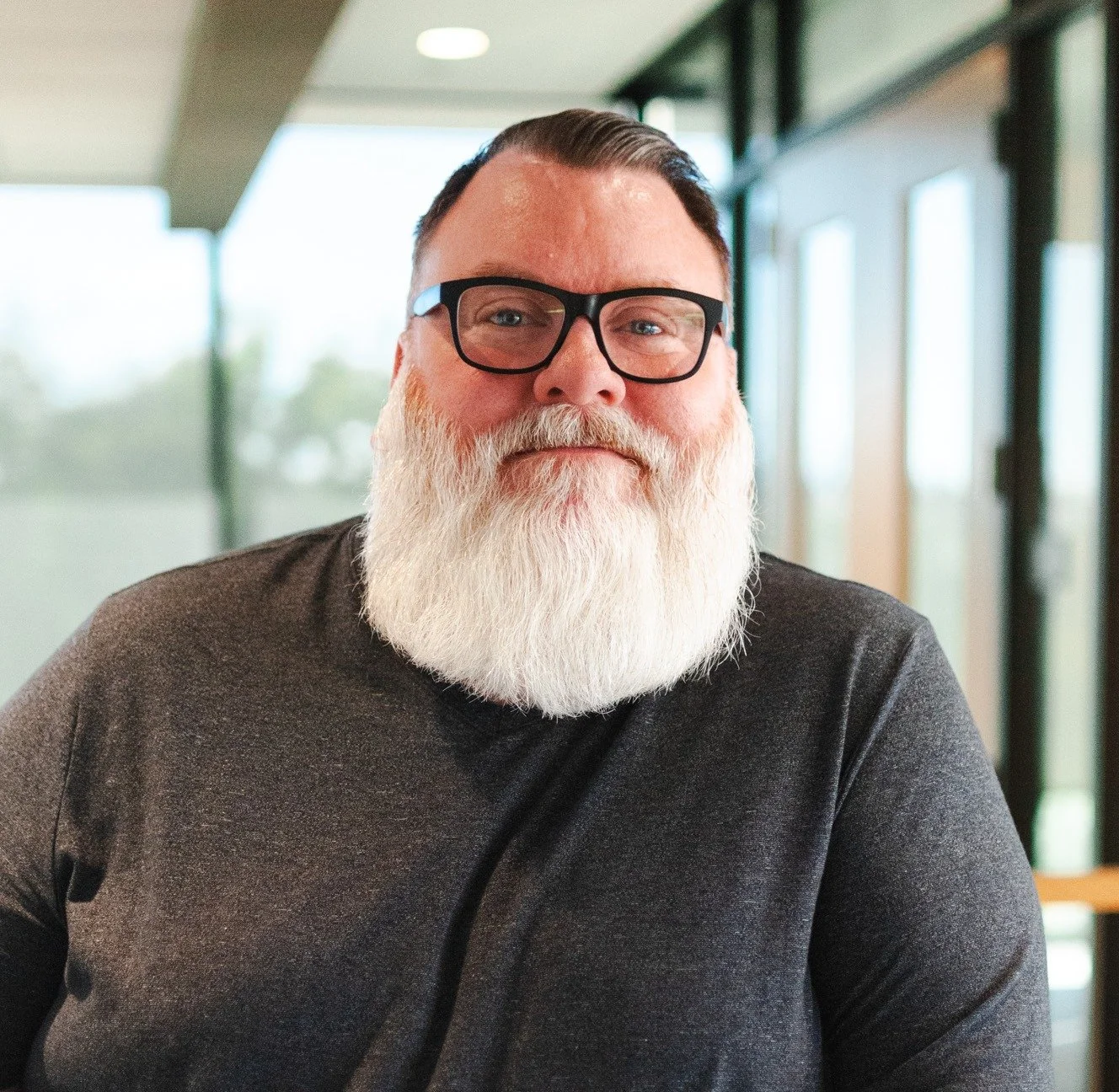 Portrait of a man with glasses and a white beard, wearing a dark gray shirt, sitting indoors near large windows.