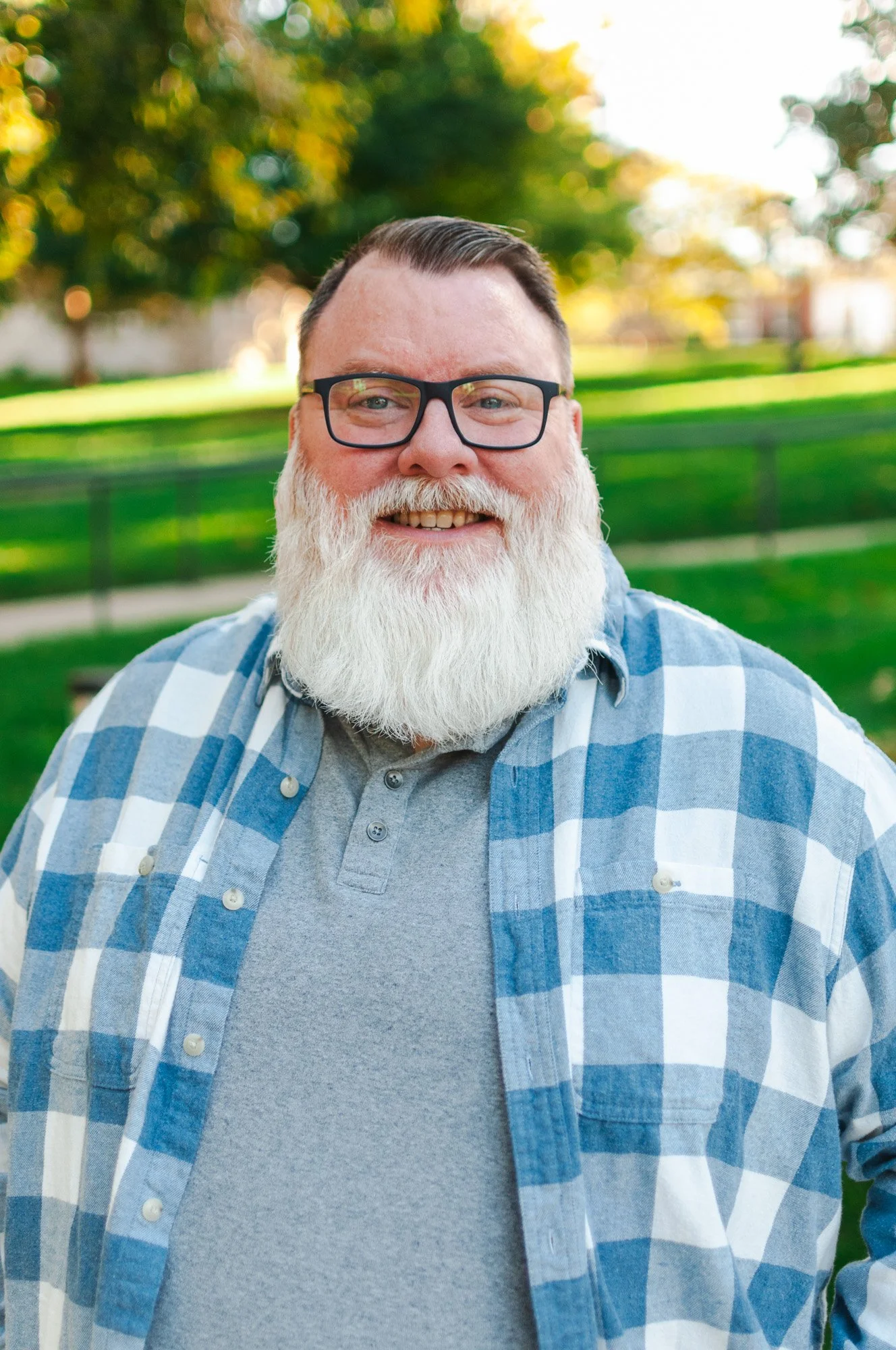 Smiling man with glasses and a full white beard, wearing a gray shirt and blue checked shirt, outdoors with green grass and trees in the background.