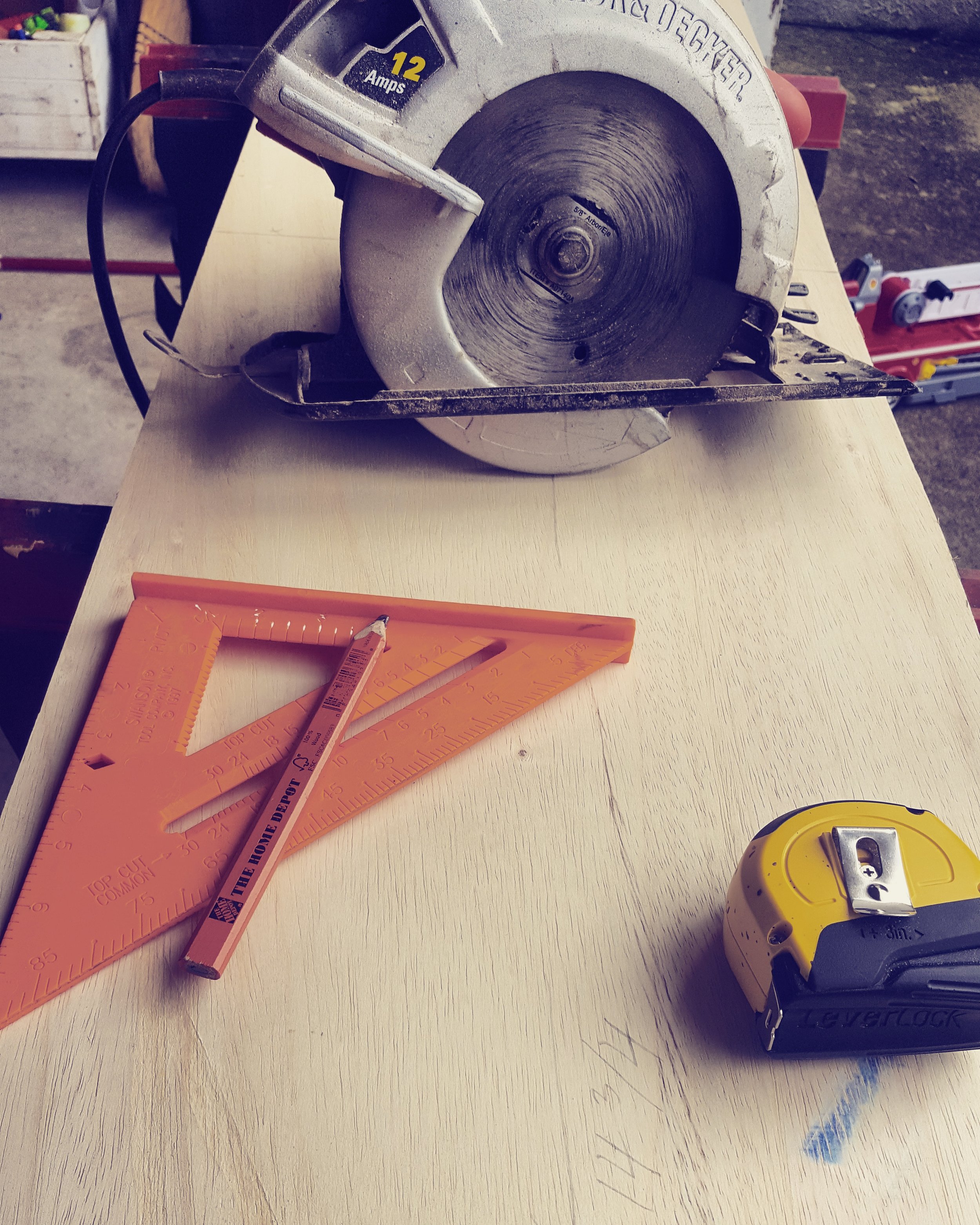 A circular saw, orange triangle rulers, a yellow measuring tape, and a yellow pencil on a light-colored wooden work surface in a workshop.