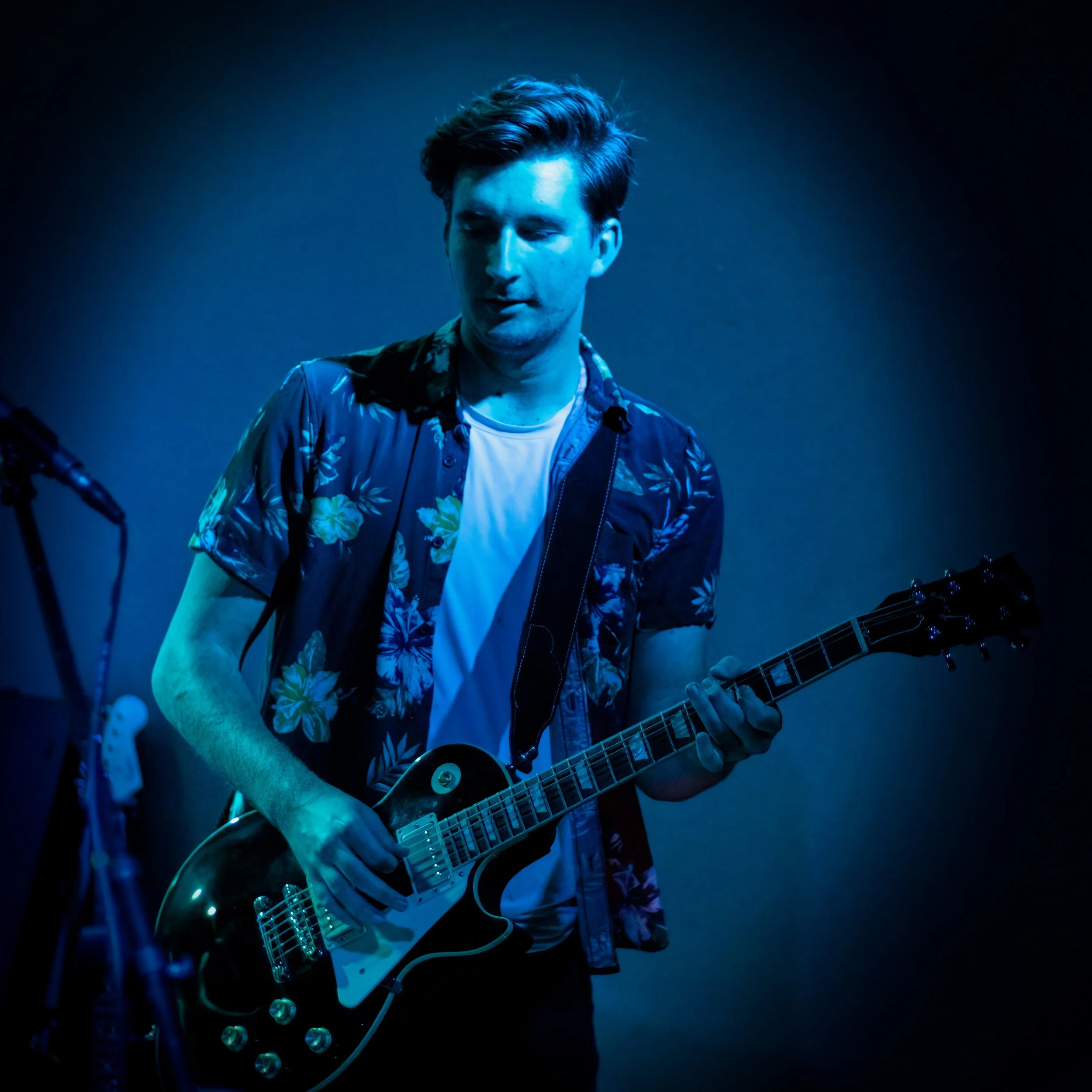 A young man with styled dark hair wearing a floral shirt over a white t-shirt, playing an electric guitar in a dark, blue-lit setting.