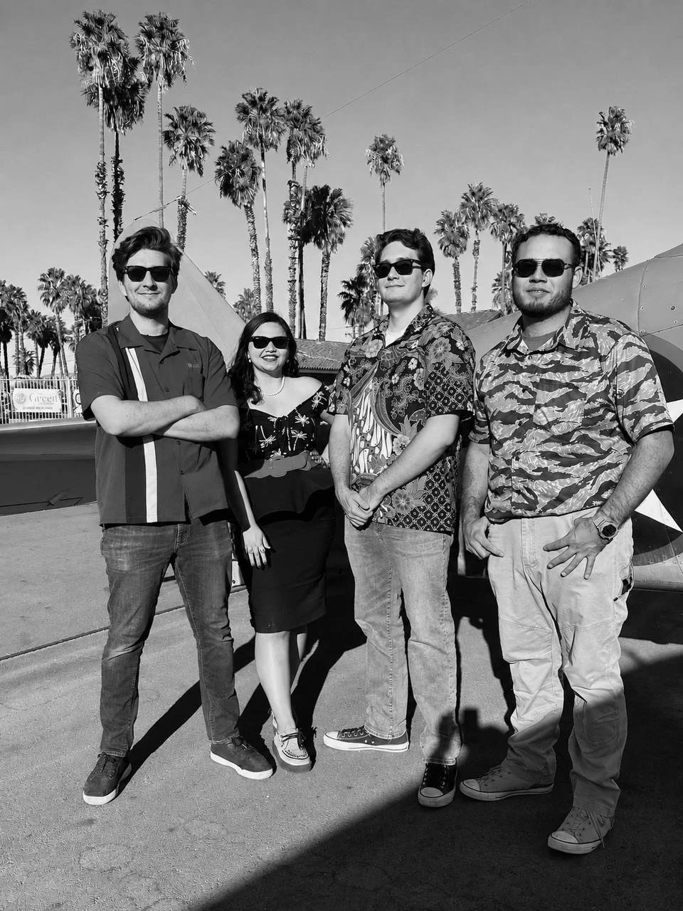 Four people standing outdoors with palm trees in the background, wearing sunglasses and casual clothing, posing for a photo.