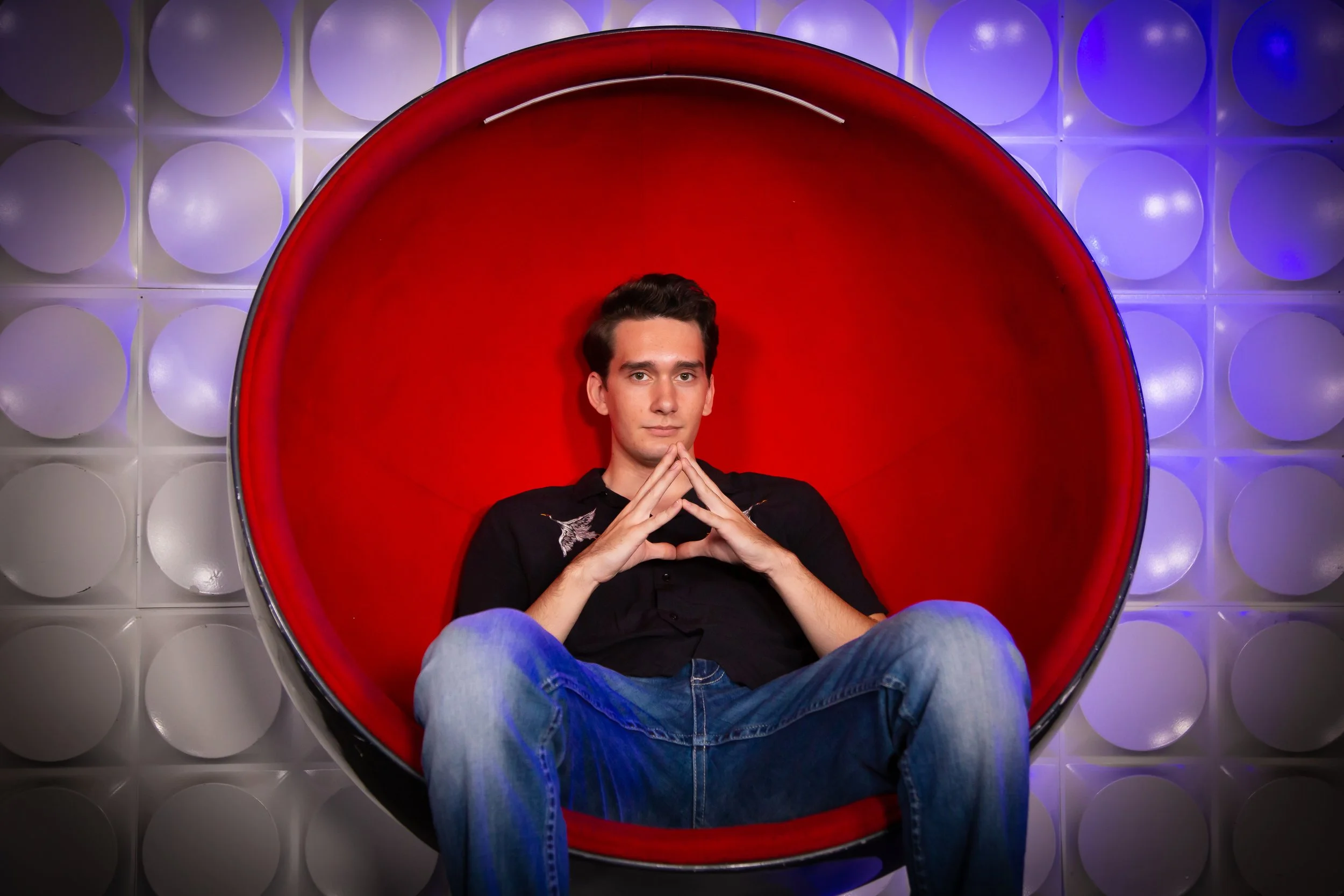 A young man with dark hair sitting inside a red, spherical chair with a high back, against a background of round, white, illuminated wall tiles, with his hands clasped in front of his face.