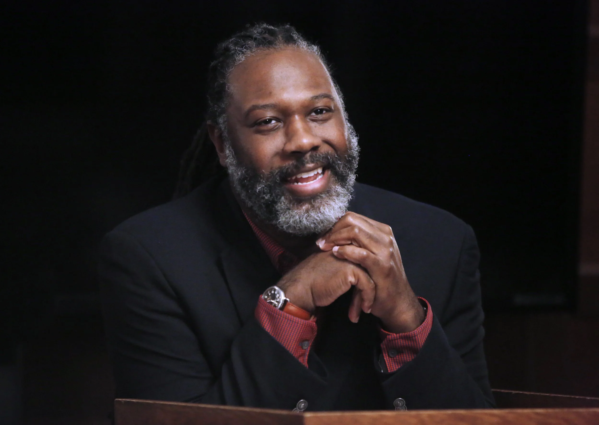 A scholar, wearing a dark blazer and a red shirt, smiling and resting his chin on his hands during an academic talk, seated against a dark background.