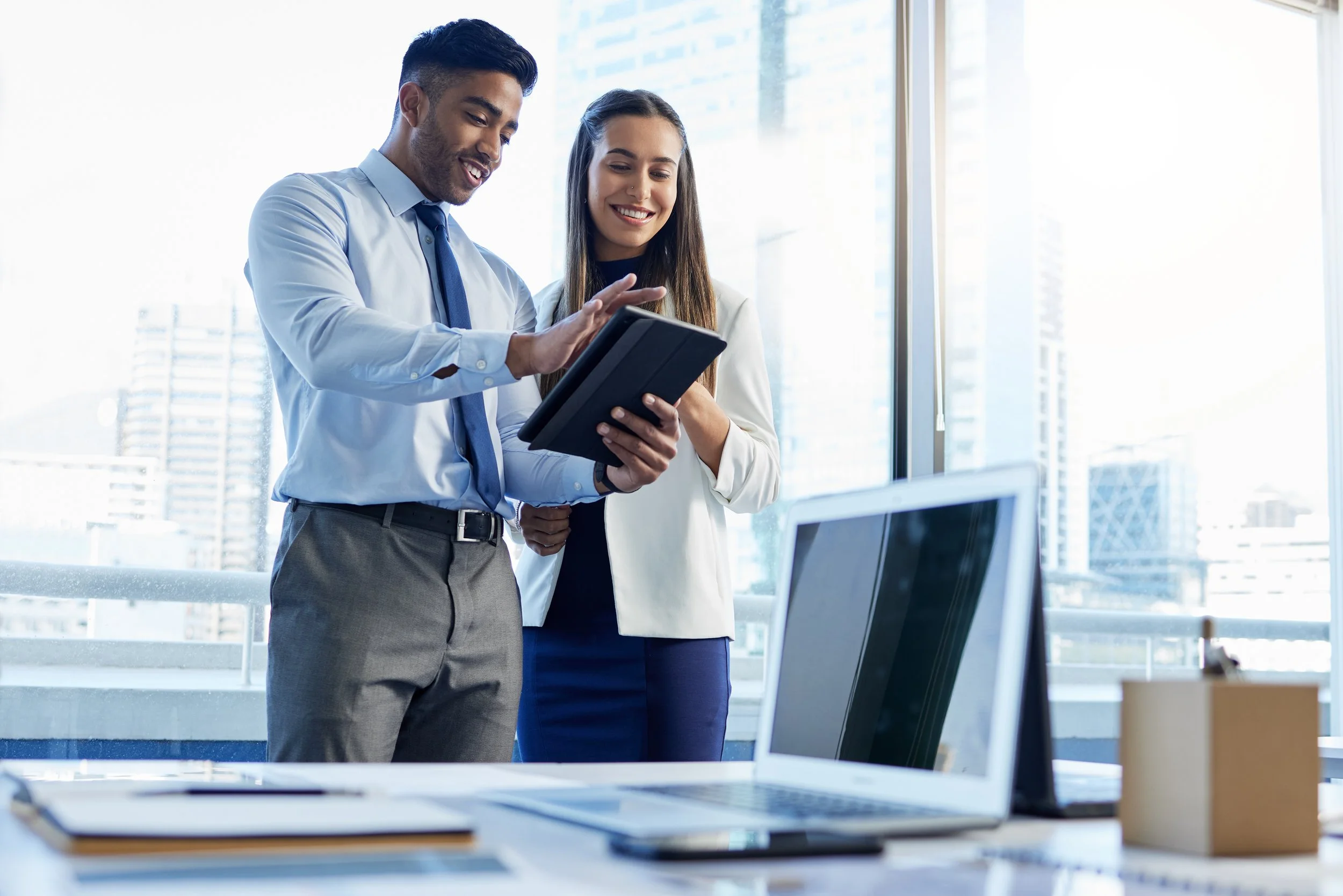 Two business professionals, a man and a woman, looking at a tablet and smiling in a modern office with large windows and cityscape view.