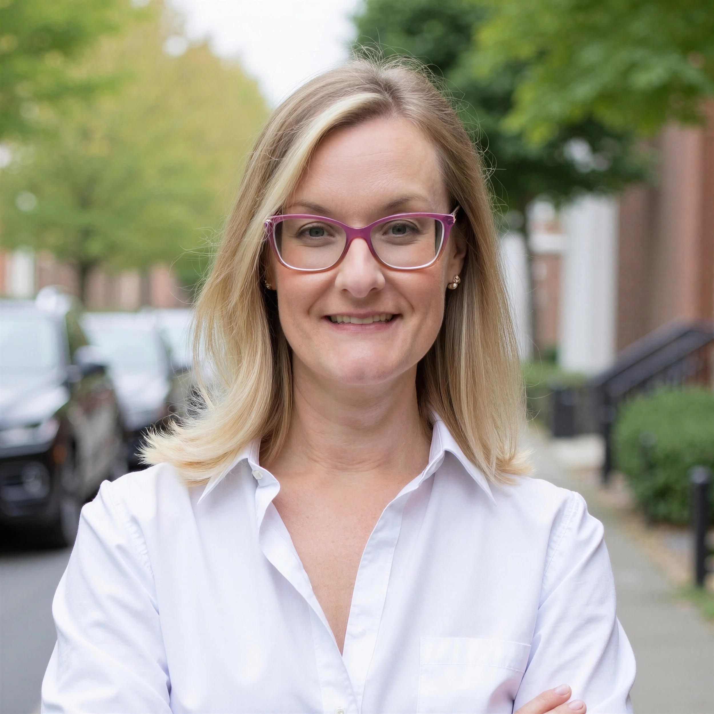 Close-up of a woman's face smiling outdoors with green trees in the background, wearing pink glasses and a white shirt.