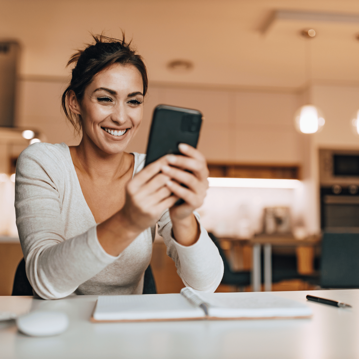 A woman with short brown hair in a beige long sleeve shirt is smiling while looking at her smartphone at a desk in a modern kitchen, looking for small business marketing assistance.
