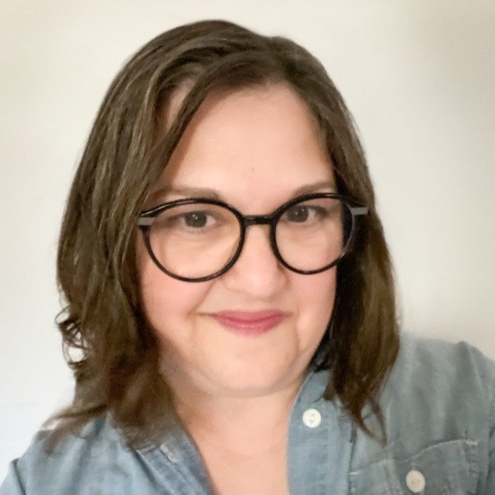 A woman with shoulder-length brown hair, wearing black-rimmed glasses and a denim shirt, smiling at the camera.
