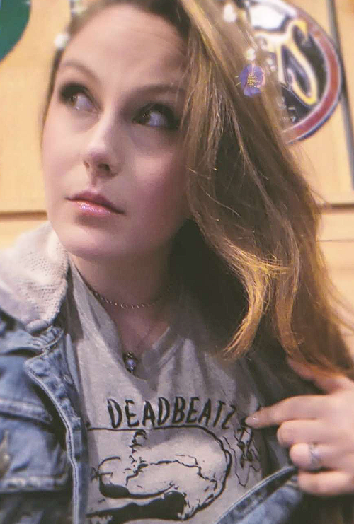 A woman with light skin and long, brown hair looking to her left while pointing to her T-shirt. She is wearing a denim jacket and a choker necklace. The background appears to be a wooden wall with a logo or sign.