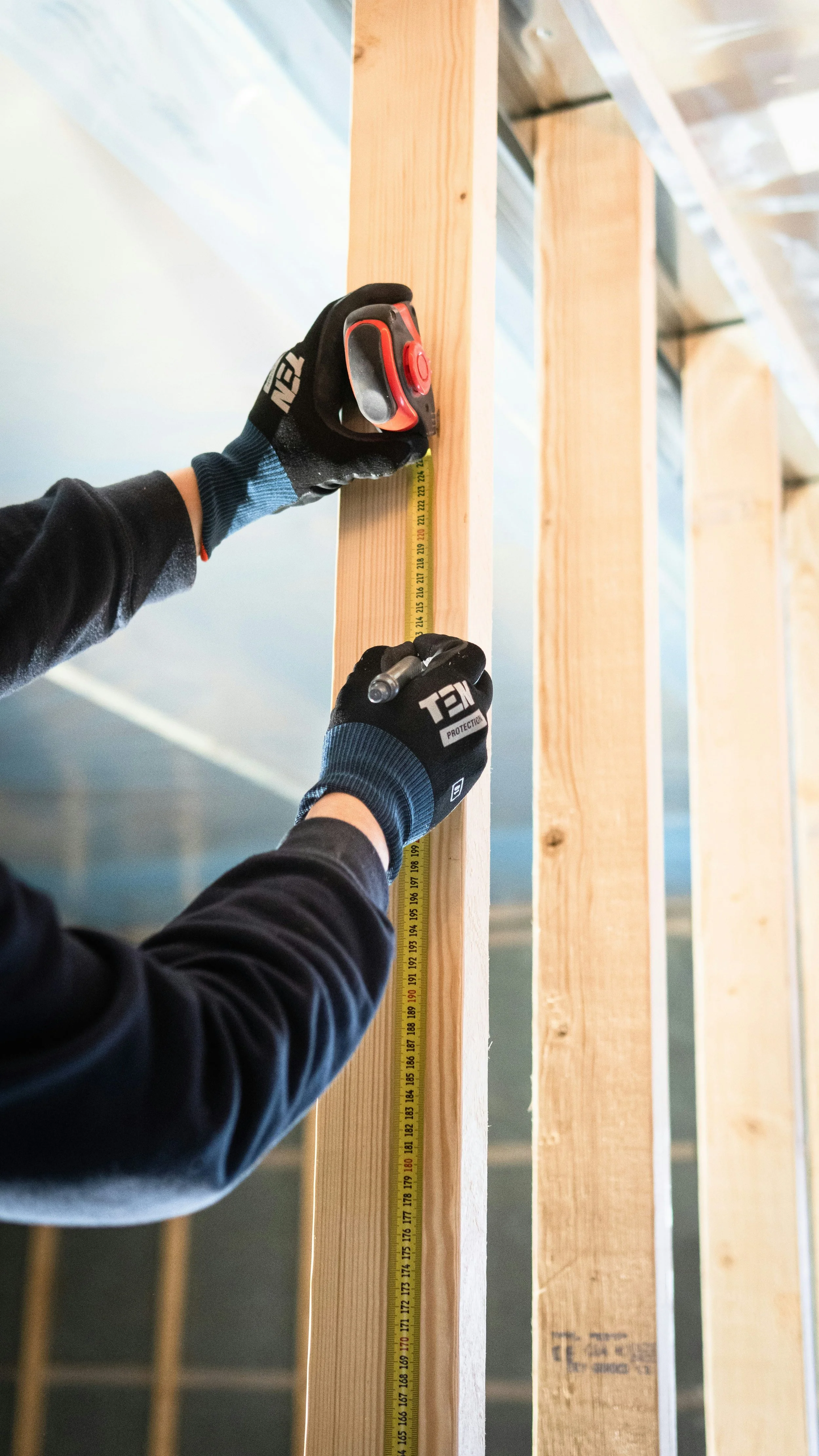A person wearing black gloves using a measuring tape and a marker to measure and mark a wooden stud for construction in a building framework.
