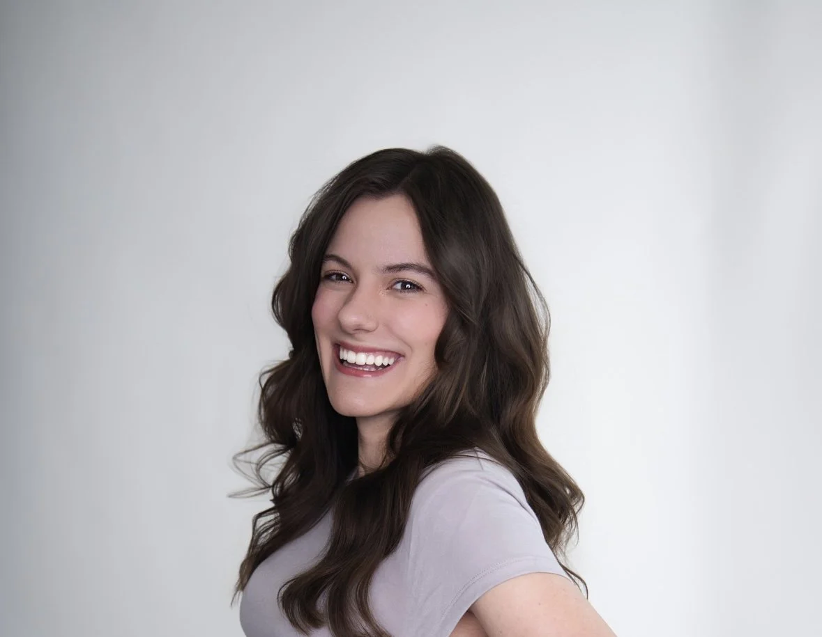 A young woman with long, wavy brown hair smiling and winking, wearing a light gray t-shirt against a plain light background.
