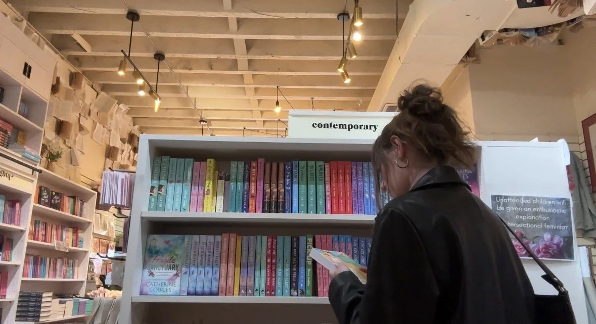 A woman wearing a black leather jacket browsing books at a bookstore, with shelves of colorful books and a sign labeled 'contemporary' in the background.
