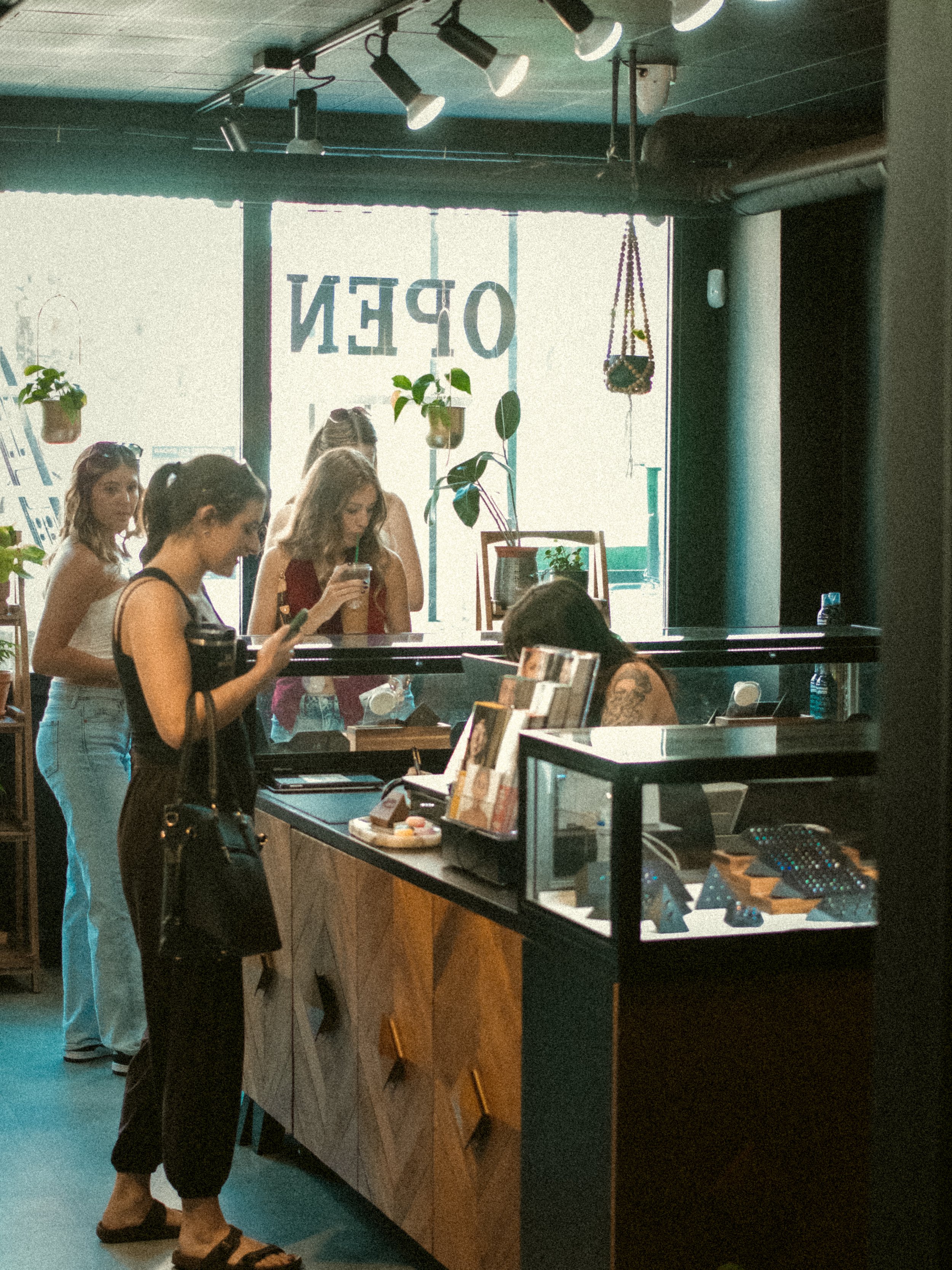 People are standing at a counter in a piercing studio, looking at the jewelry and completing paperwork. The setting includes a large window with the word 'OPEN' visible, indoor plants, and track lighting on the ceiling.