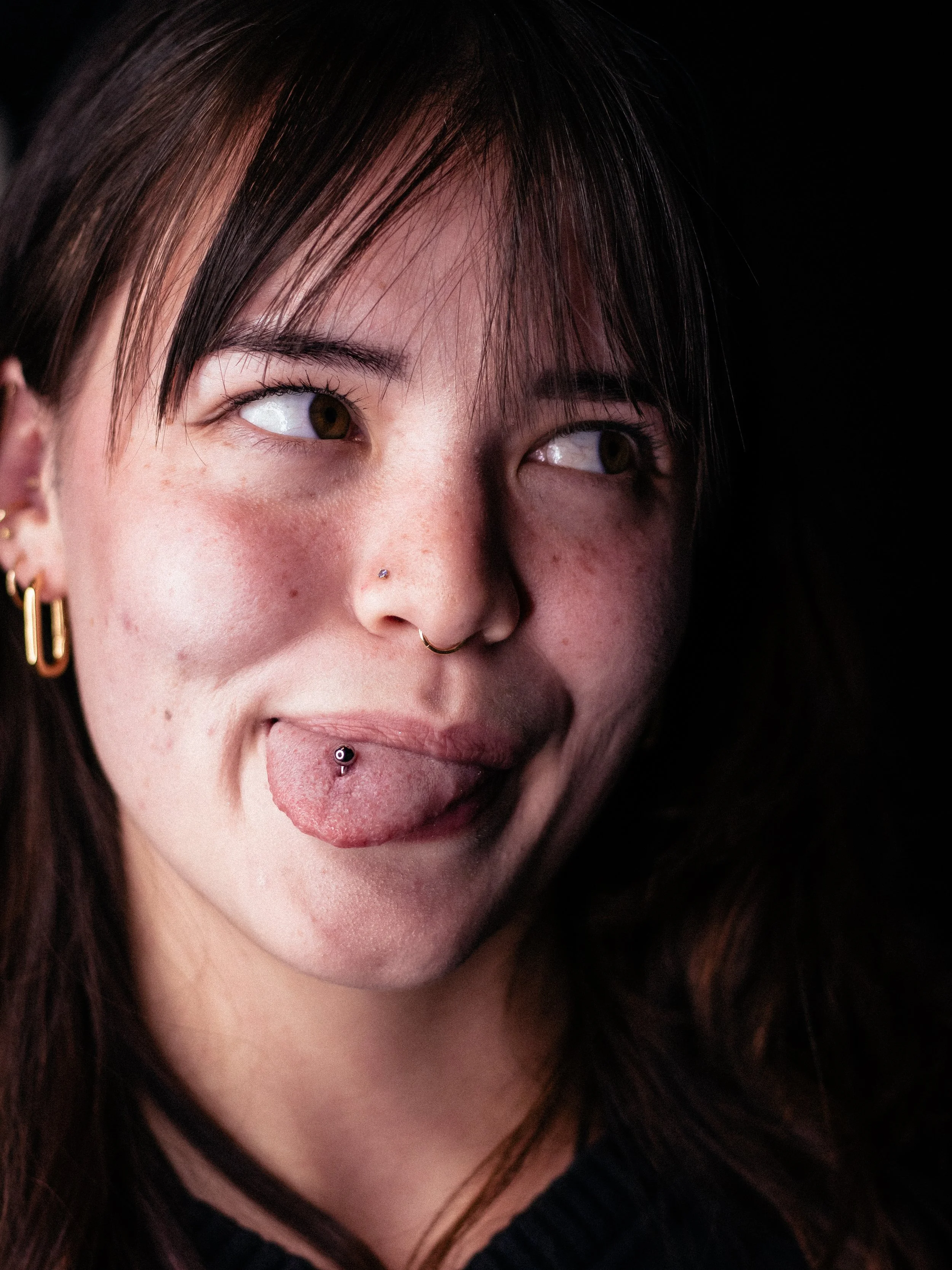 Close-up of a young woman with brown eyes, dark brown hair, multiple earrings, nose piercings, and sticking out her tongue.
