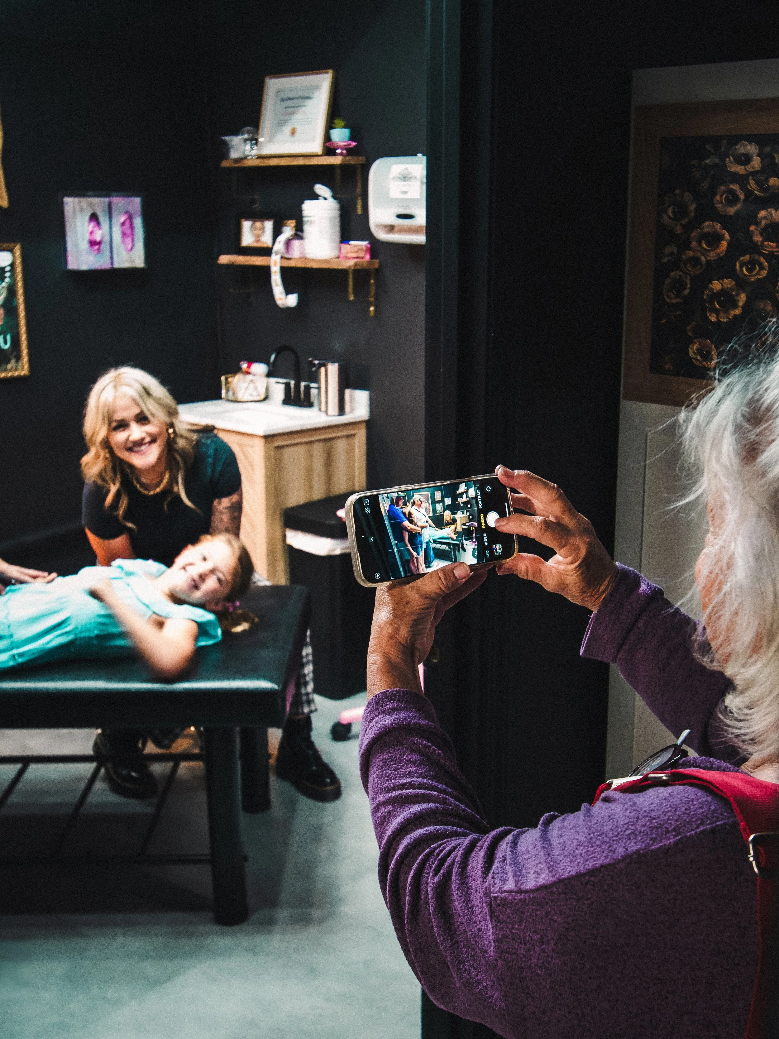 A woman with white hair and a purple sweater is taking a picture with her phone of a woman with blonde hair and a dark shirt who is smiling and leaning over a young girl with a light blue dress lying on a black table. The scene is in a room with dark walls and some framed pictures and shelves with personal items.