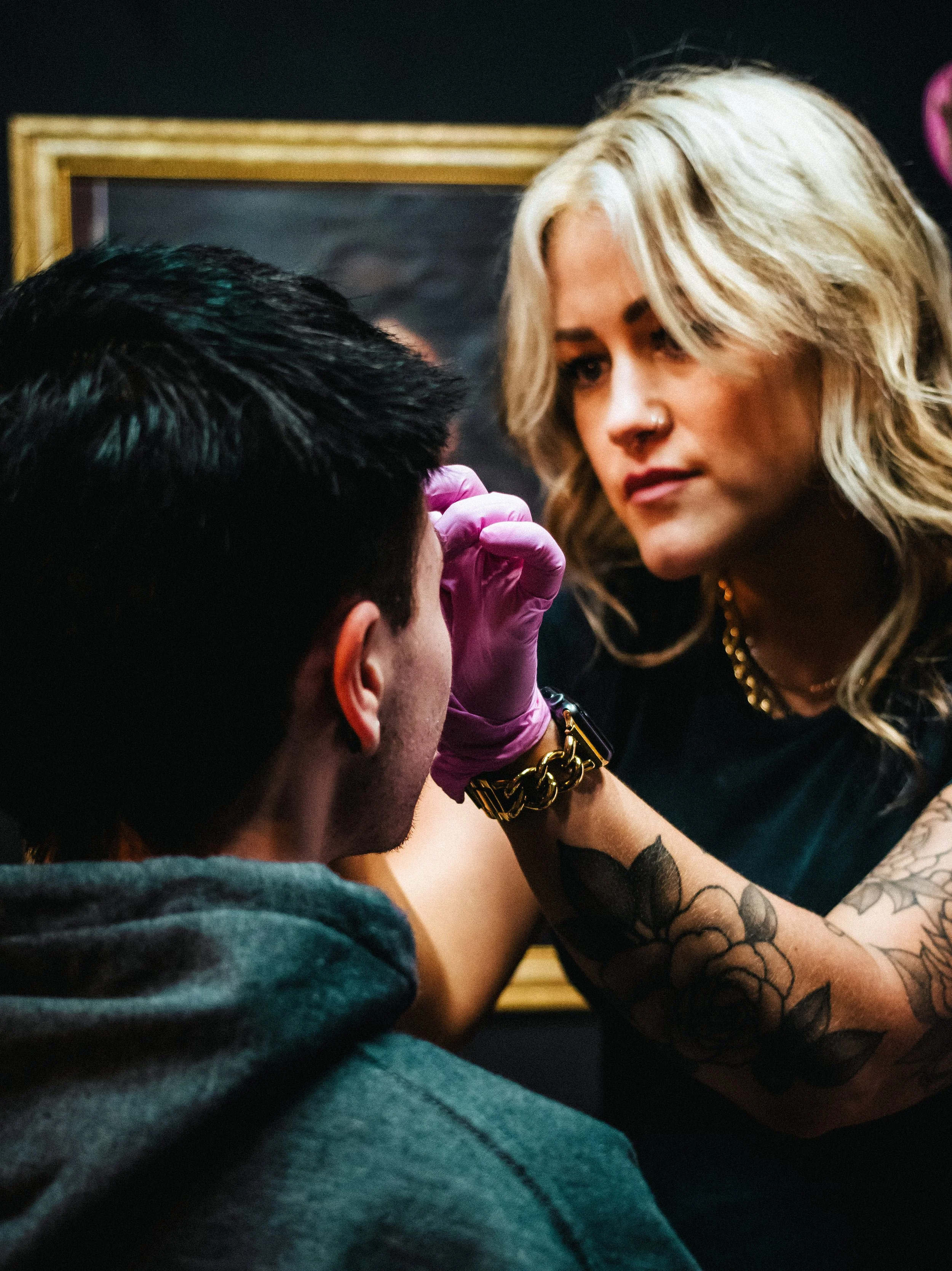 A woman with blonde hair and tattoos is piercing a man's eyebrow as he sits still, wearing a dark shirt and a purple glove.