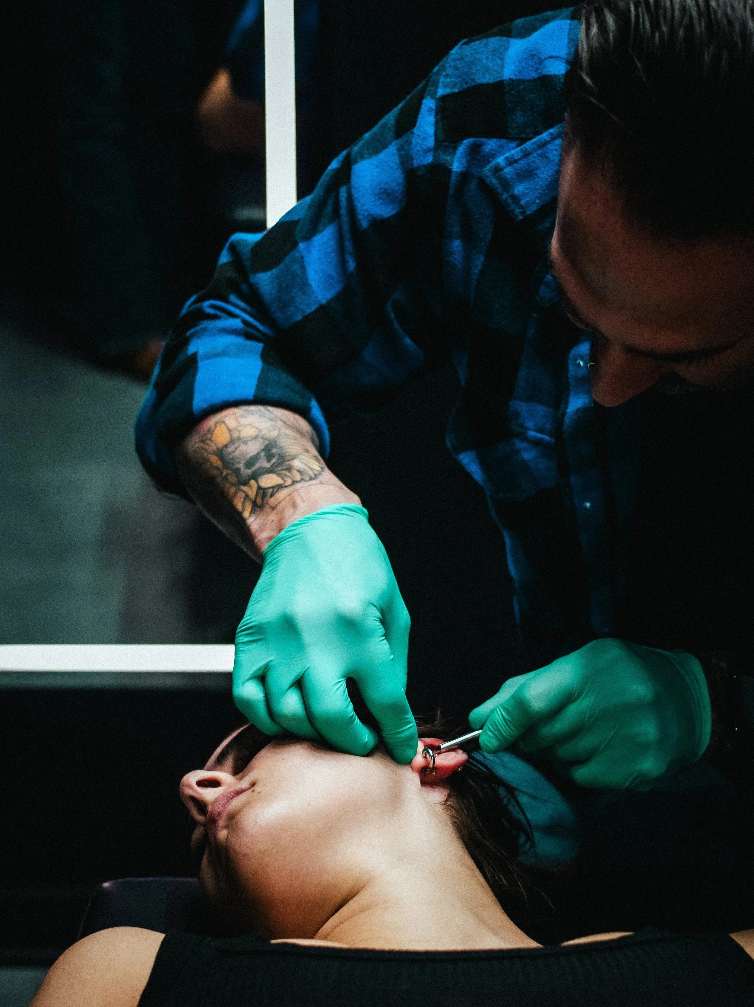 A person lying down with eyes closed as a body piercer performs a piercing on their ear. The piercer wears green gloves and a blue checkered shirt.