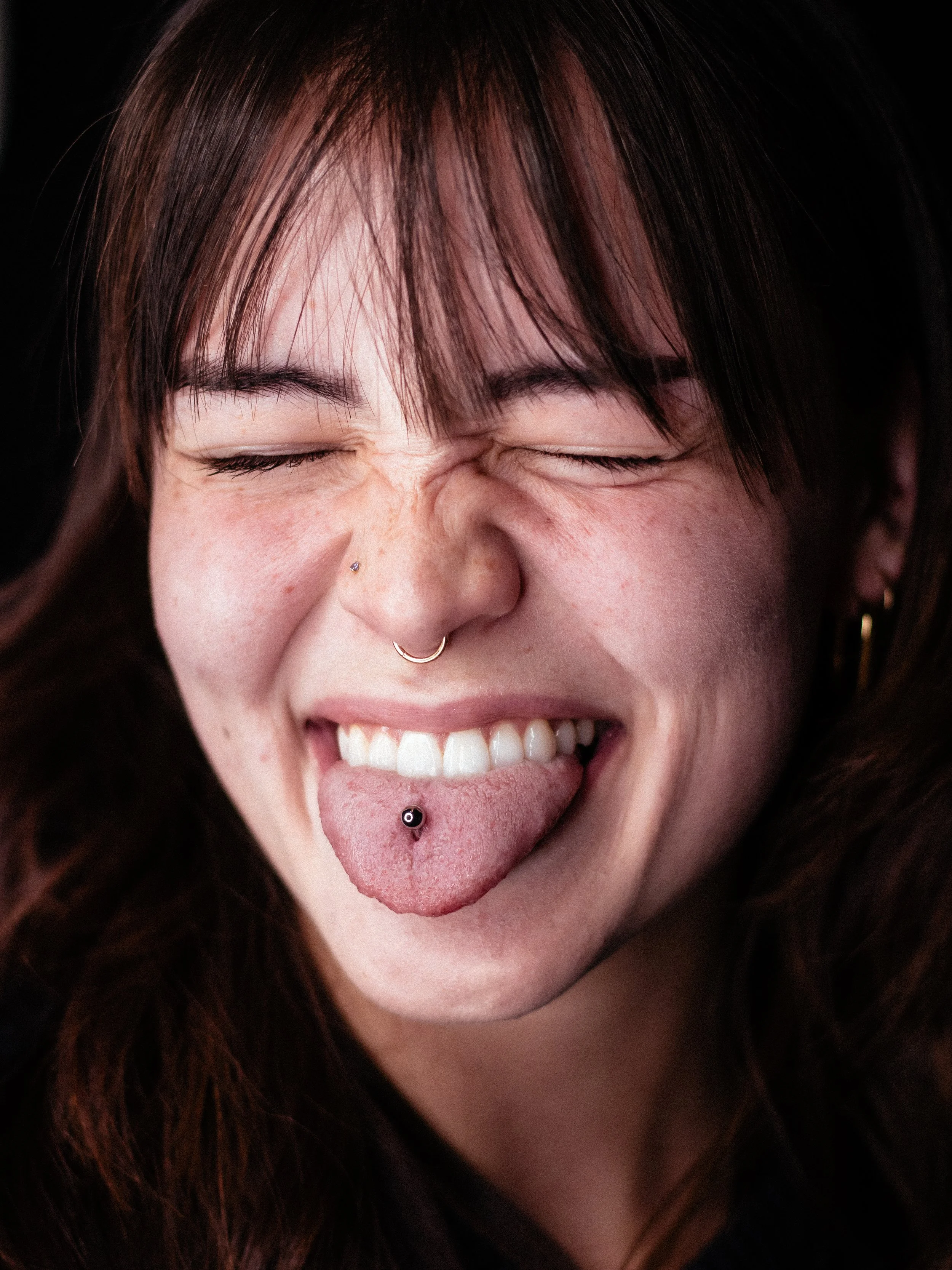 Close-up of a person with brown hair, closed eyes, and tongue sticking out, showing a tongue piercing.
