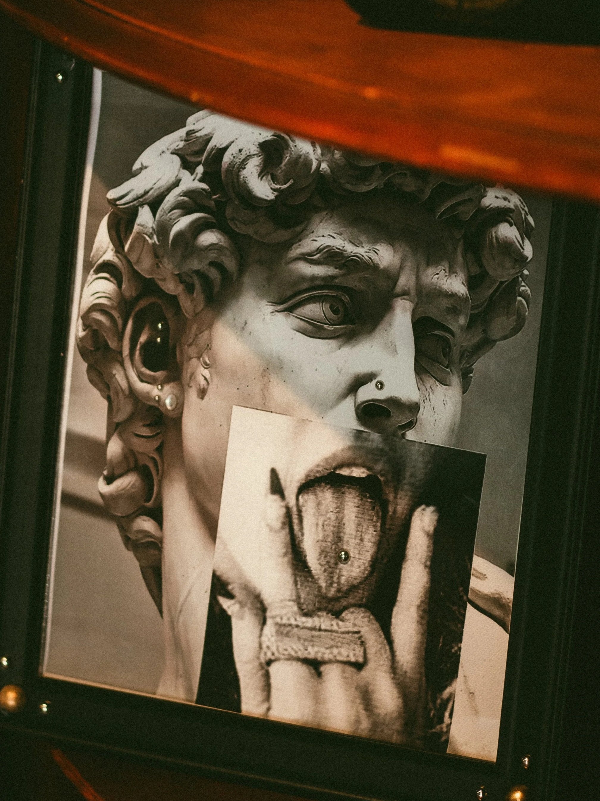 A photograph of a classical marble sculpture of a woman's face with curly hair, shown from a side angle, partially obscured by a reddish-orange surface in the foreground.