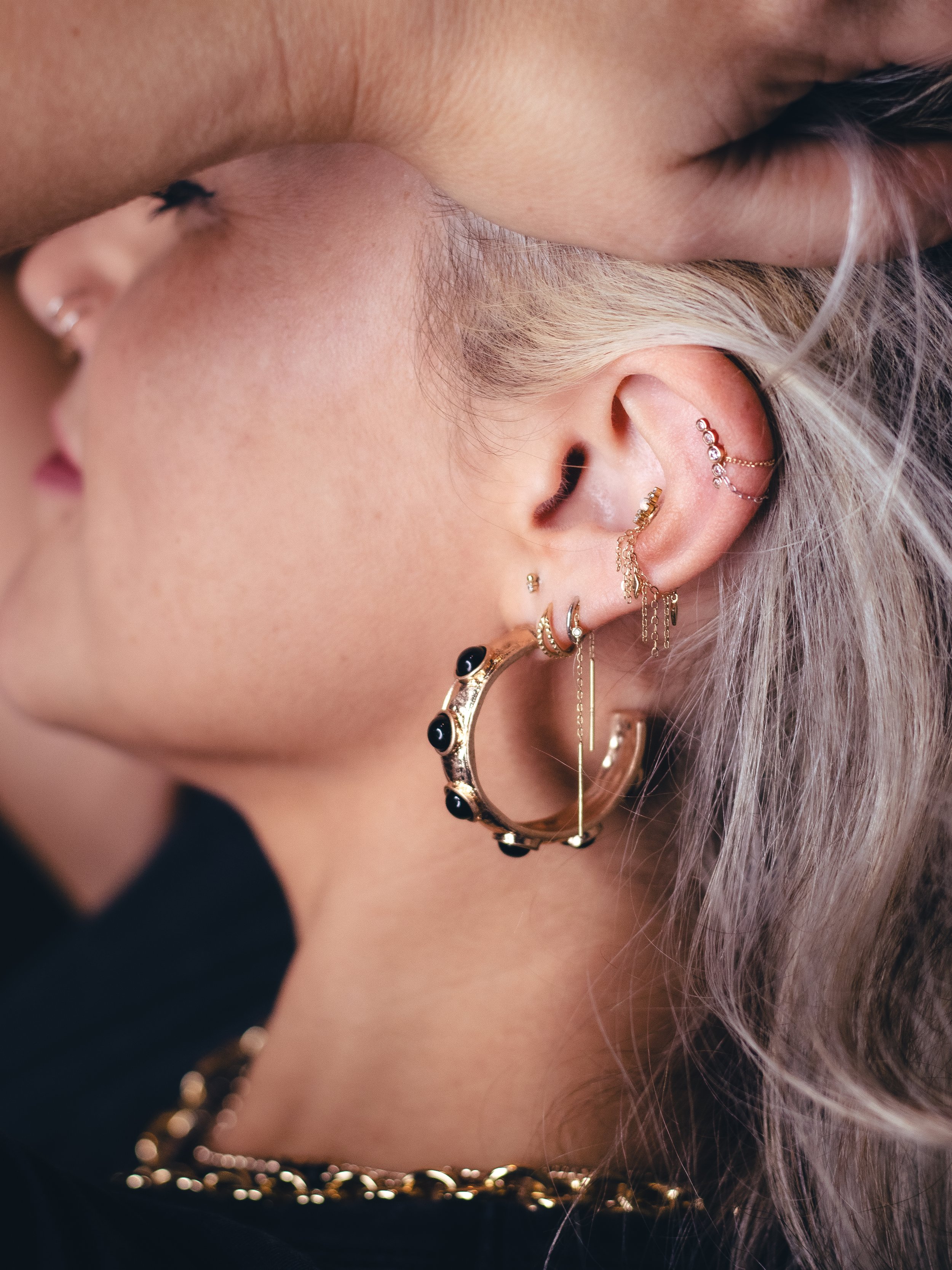 Close-up of a person's ear decorated with multiple gold earrings and a three-layered pearl and gold chain necklace, with long, platinum blonde hair.