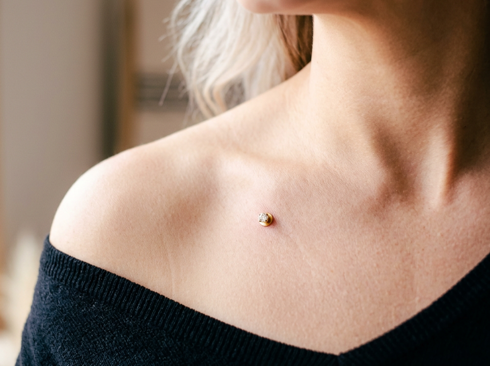 Close-up of a woman's shoulder and collarbone, showing a small, round gold and diamond surface anchor piercing on her shoulder. She is wearing a black off-the-shoulder top.