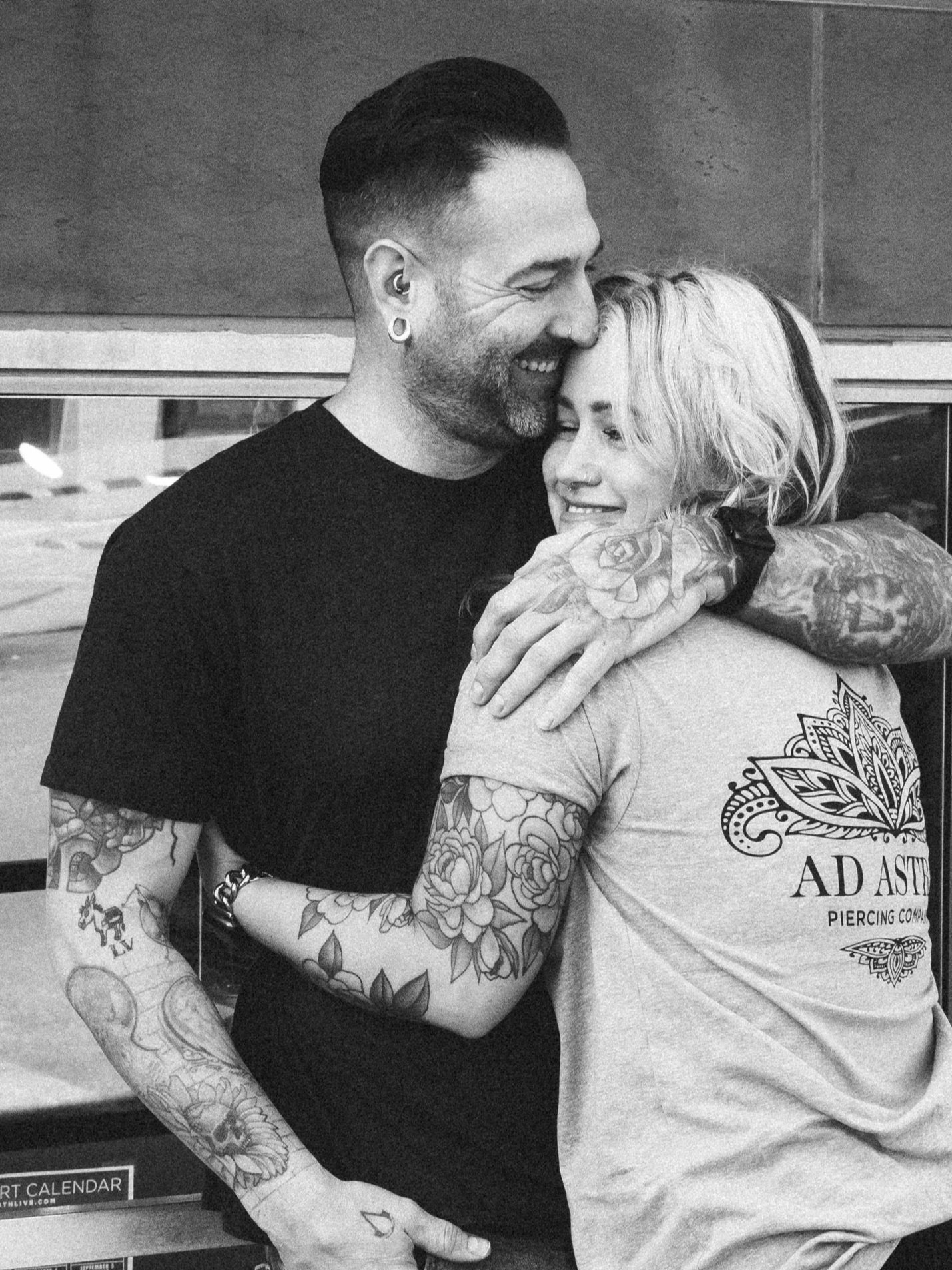 A tattooed man and a woman embrace and smile, with the man’s face close to the woman’s forehead. They are smiling and seem to be happy.