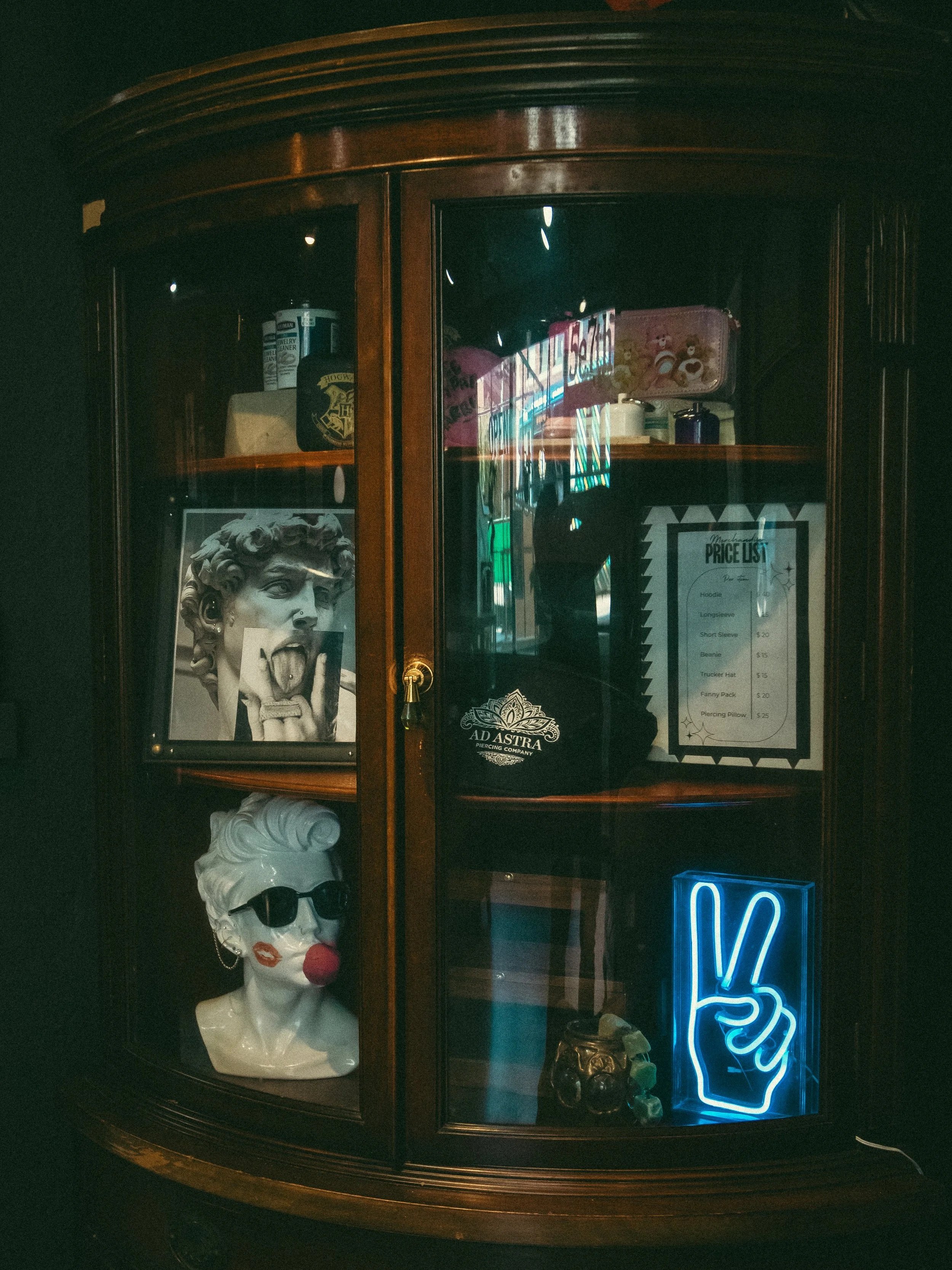 A wooden display cabinet with various items inside, including a gray bust of a woman with sunglasses, a bartender sign, a colorfully labeled tin box, a price list, and a neon blue thumbs-up sign at the bottom.