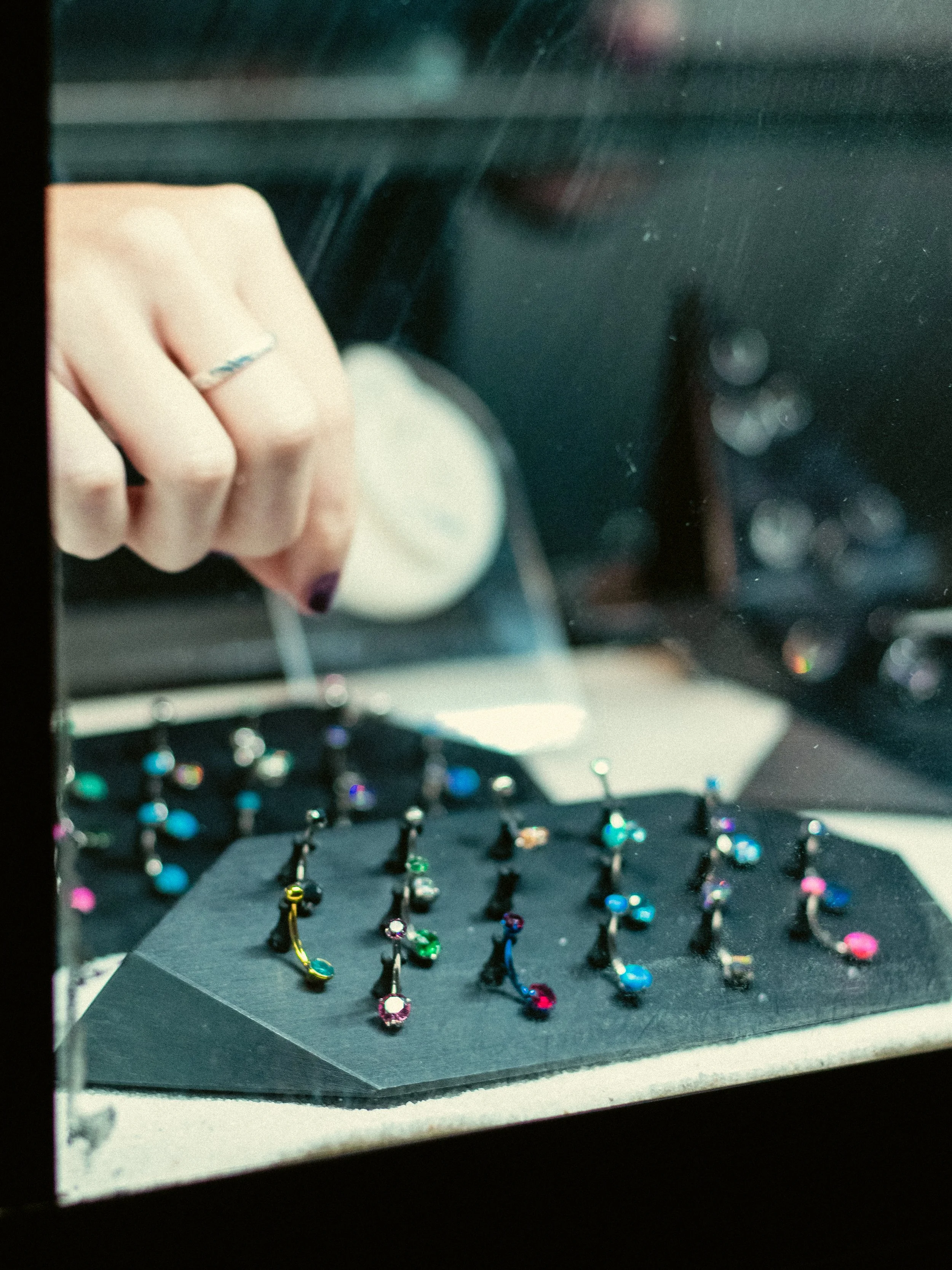 Close-up of a display case with colorful rings arranged on black foam, a hand with painted nails and a silver ring is reaching inside, and blurred multicolored lights in the background.