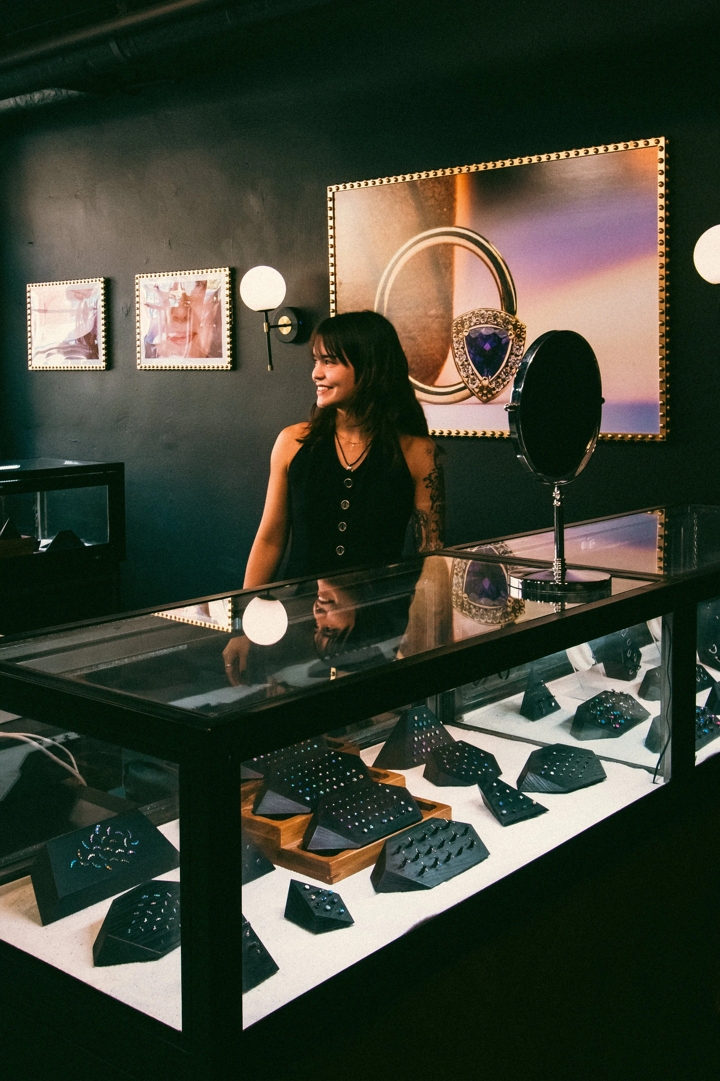 A woman shopping for jewelry at a display table with various rings and earrings in a jewelry store. There are pictures of jewelry on the dark wall behind her, including a large picture of a ring with a large gemstone.