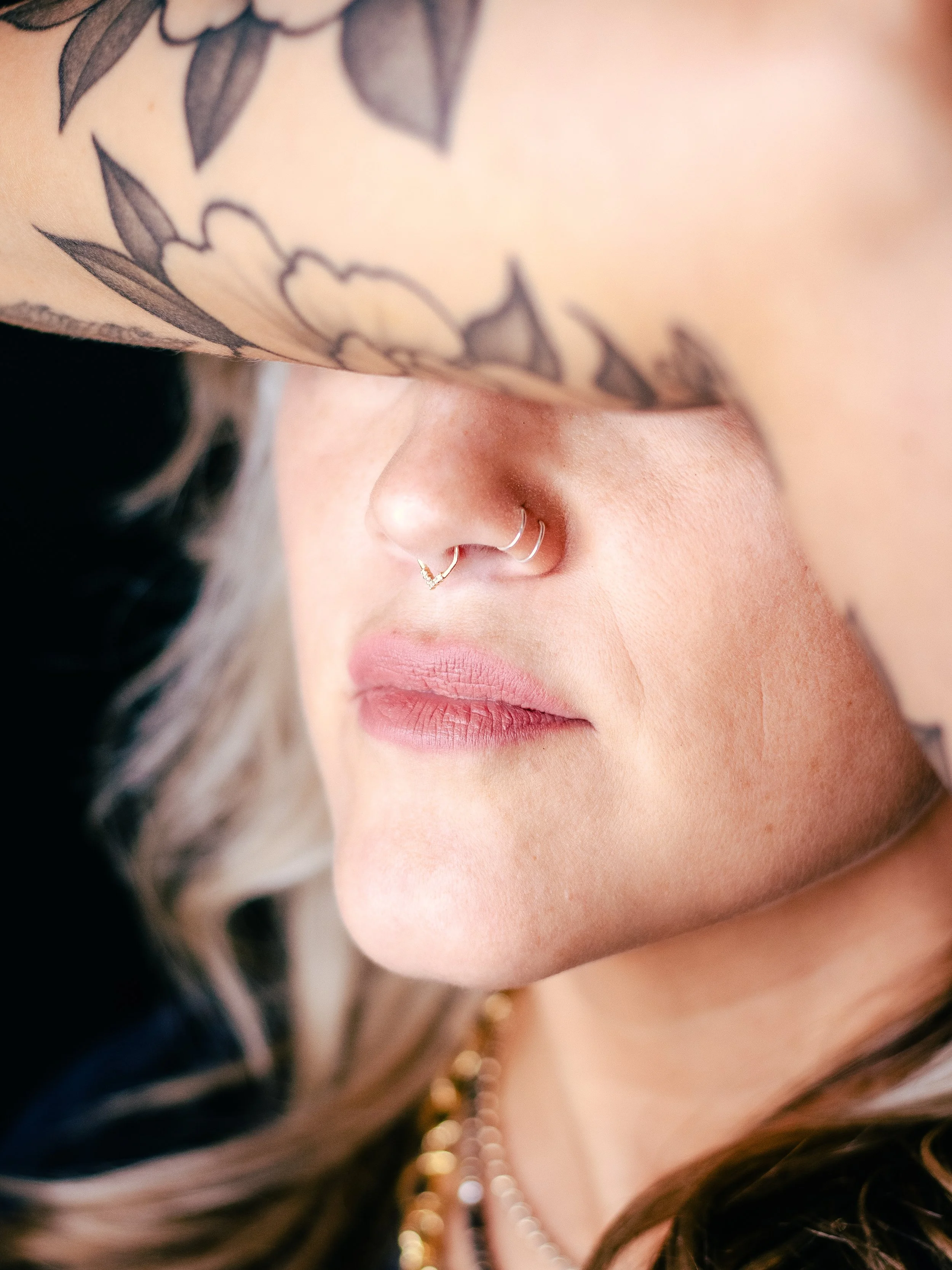 Close-up of a person's face showing a nose with multiple nose rings and a septum piercing, partially covered by her arm with floral tattoos. The person has light skin and dark hair.
