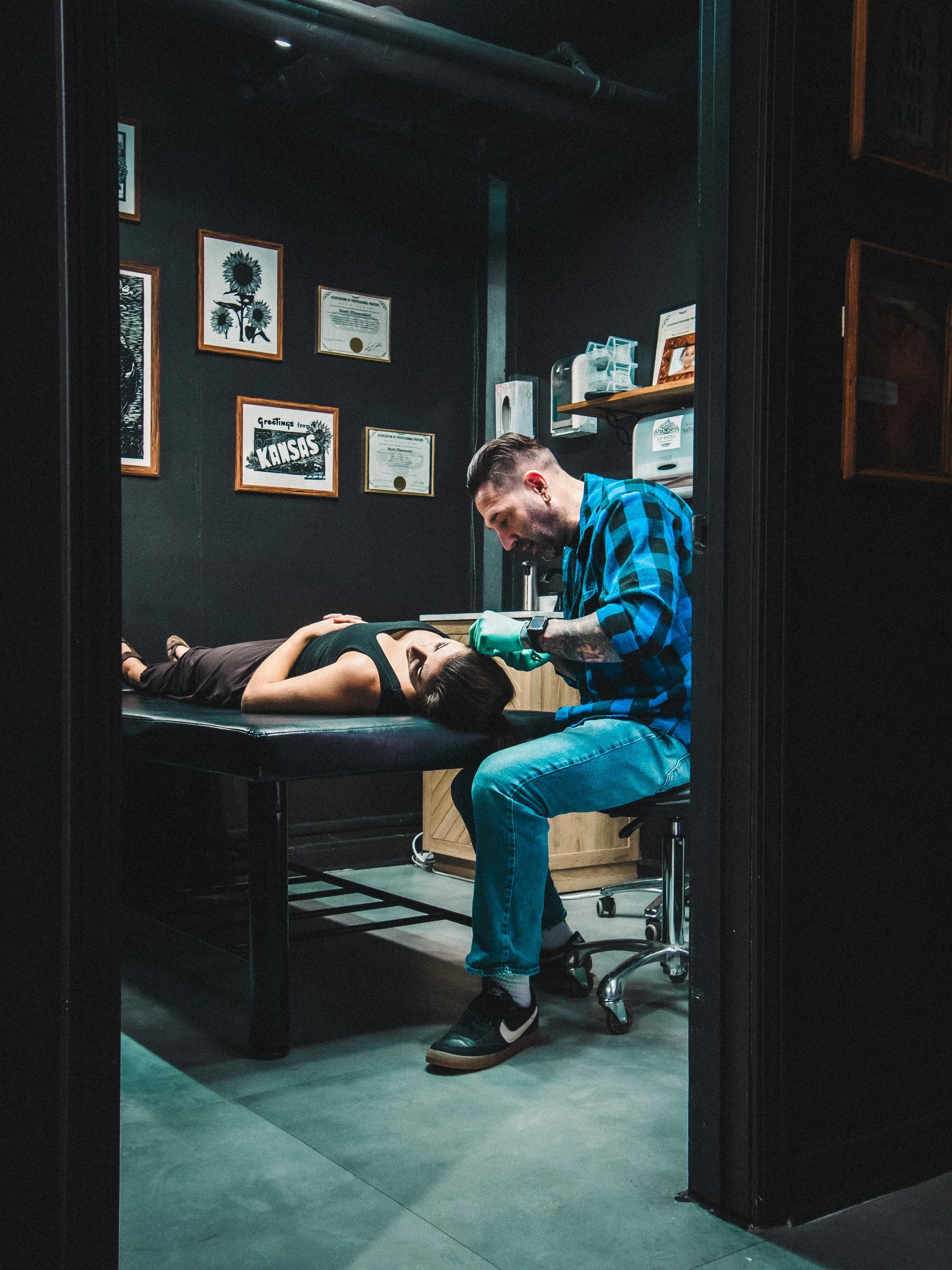 Licensed professional piercer with short hair and ear piercings piercing a woman lying on a table in a piercing shop in Kansas.