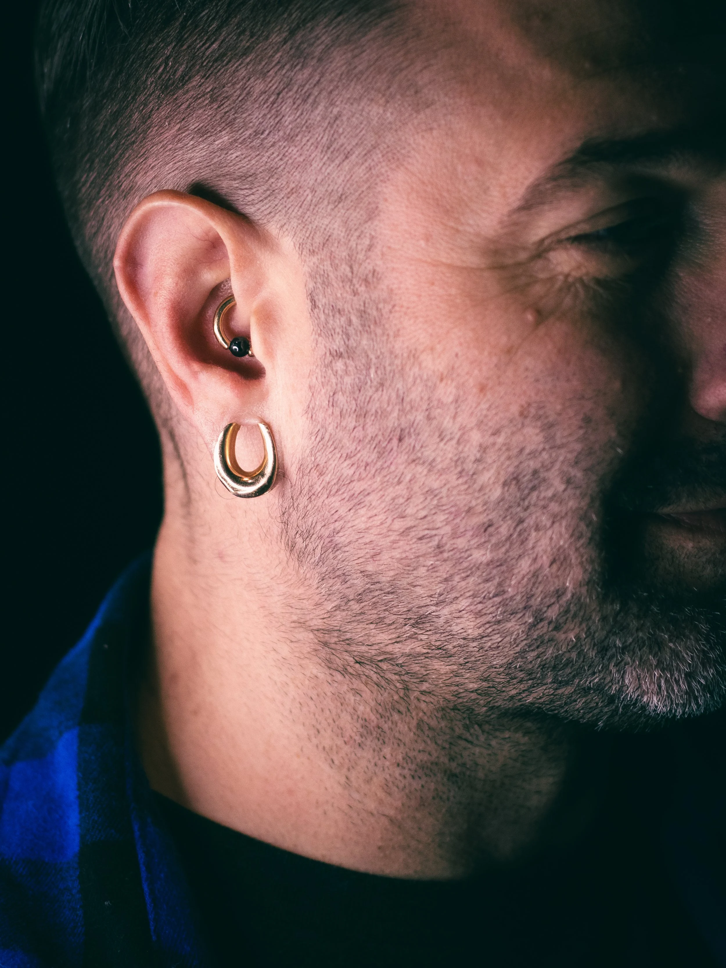 Close-up of a person's ear with multiple earrings, including a small black stud and a gold hoop, alongside short hair and part of a beard.