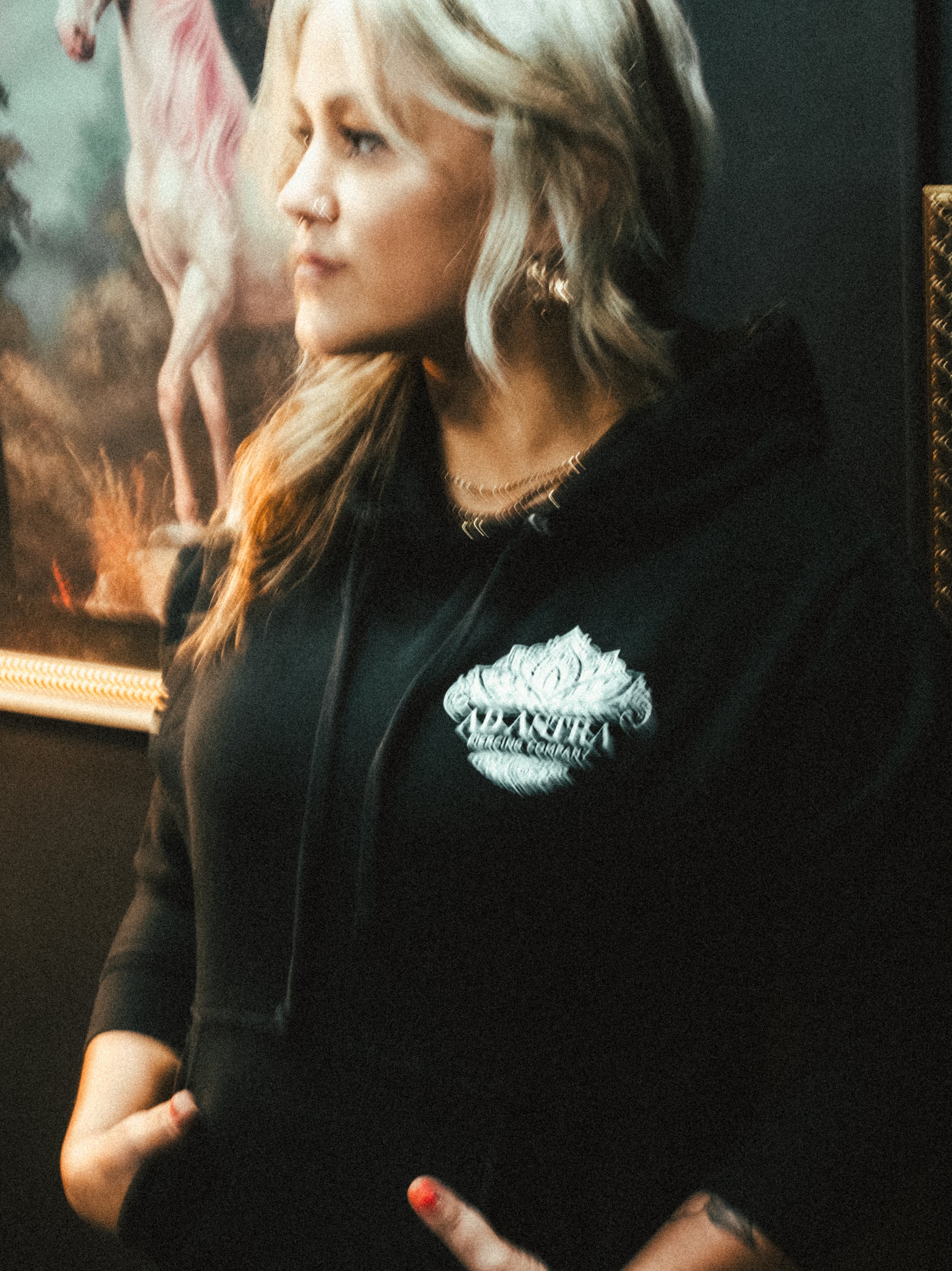 A woman with blonde hair and a nose piercing wearing a black hoodie with a white logo and silver jewelry, standing in front of a colorful, framed poster or picture.