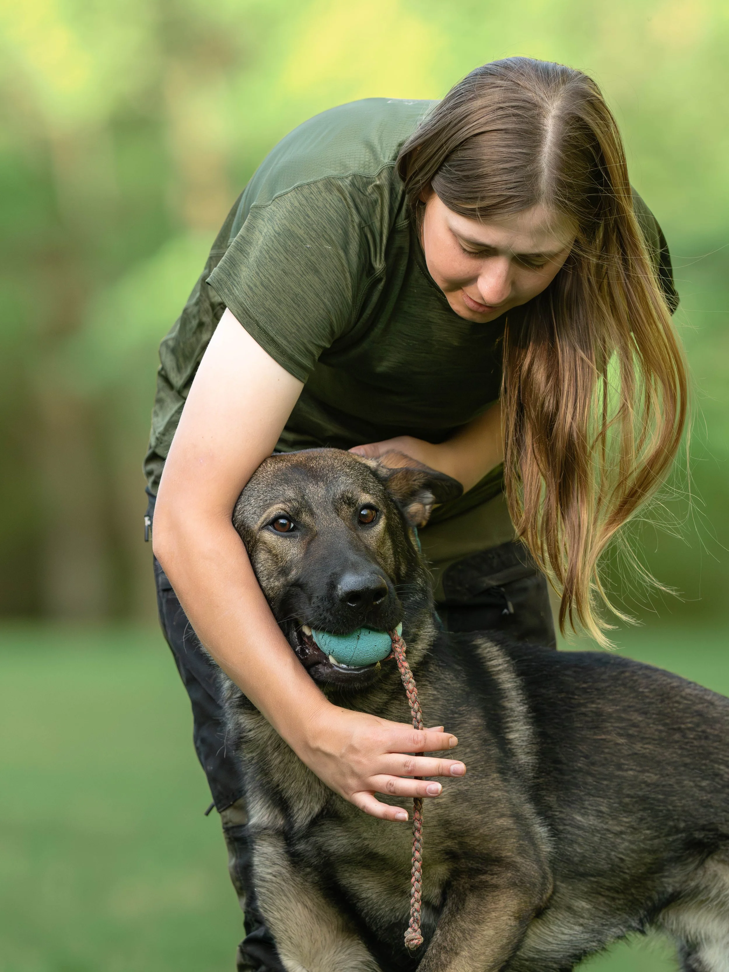 A woman with long brown hair and a green shirt hugging a large German Shepherd dog with a blue ball in its mouth outdoors in a grassy area.