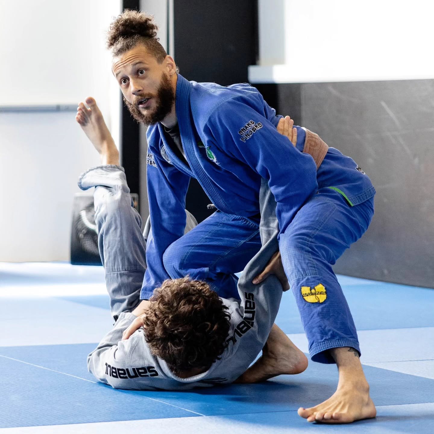 Two individuals practicing Brazilian Jiu-Jitsu on a blue mat. One person in a blue gi is on top, controlling the person in a gray gi on the ground.
