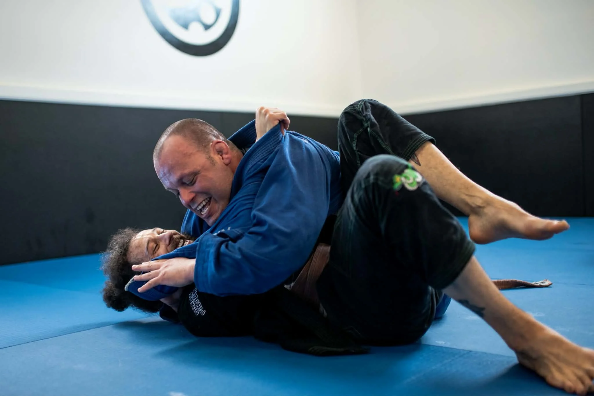 Two martial artists practicing Brazilian Jiu-Jitsu on a blue mat, engaged in a grappling position with one person on top and the other on the bottom, smiling and dressed in traditional uniforms.
