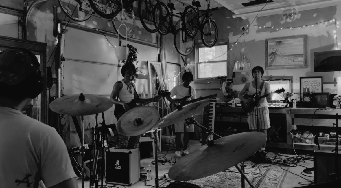 A band of five musicians playing instruments in a cozy, decorated room with bicycles hanging from the ceiling and string lights. The musicians include a drummer, guitarist, keyboardist, bass player, and singer with an acoustic guitar.