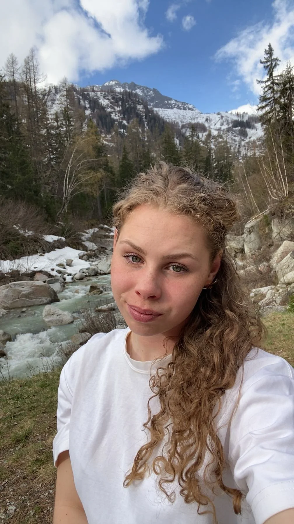 Portret van een jonge vrouw met lang, gekruld haar voor een berglandschap met een snelstromende rivier, dennenbomen en bergen met sneeuw op de achtergrond.